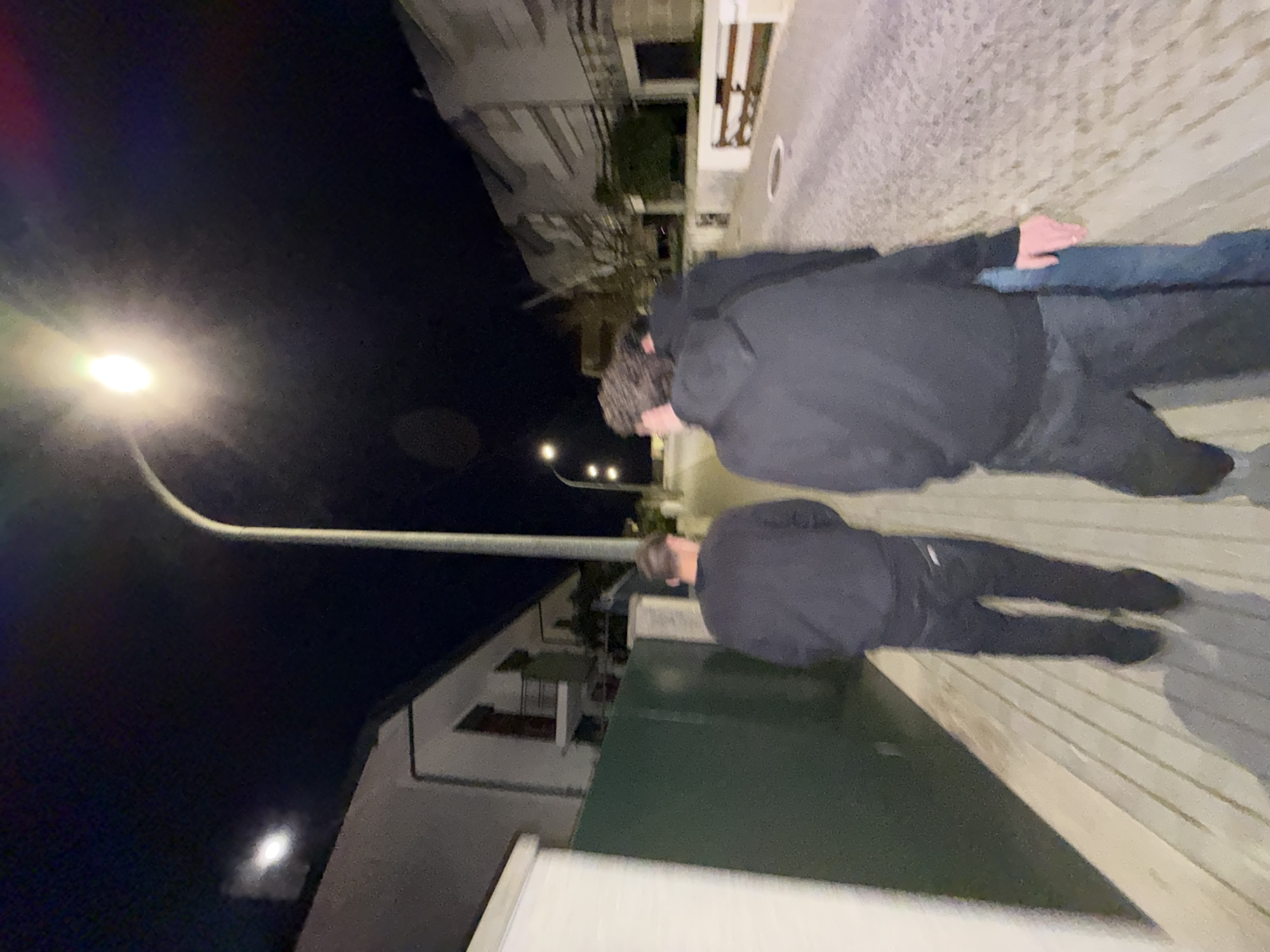 Two people are walking on a paved street in Vila Real, Portugal, at night. The street is illuminated by a streetlight. Buildings can be seen on both sides of the street. The person closest to the viewer has brown curly hair and is wearing a black hoodie, dark jeans, and black shoes. The other person is wearing a black hoodie, dark jeans, and black shoes.