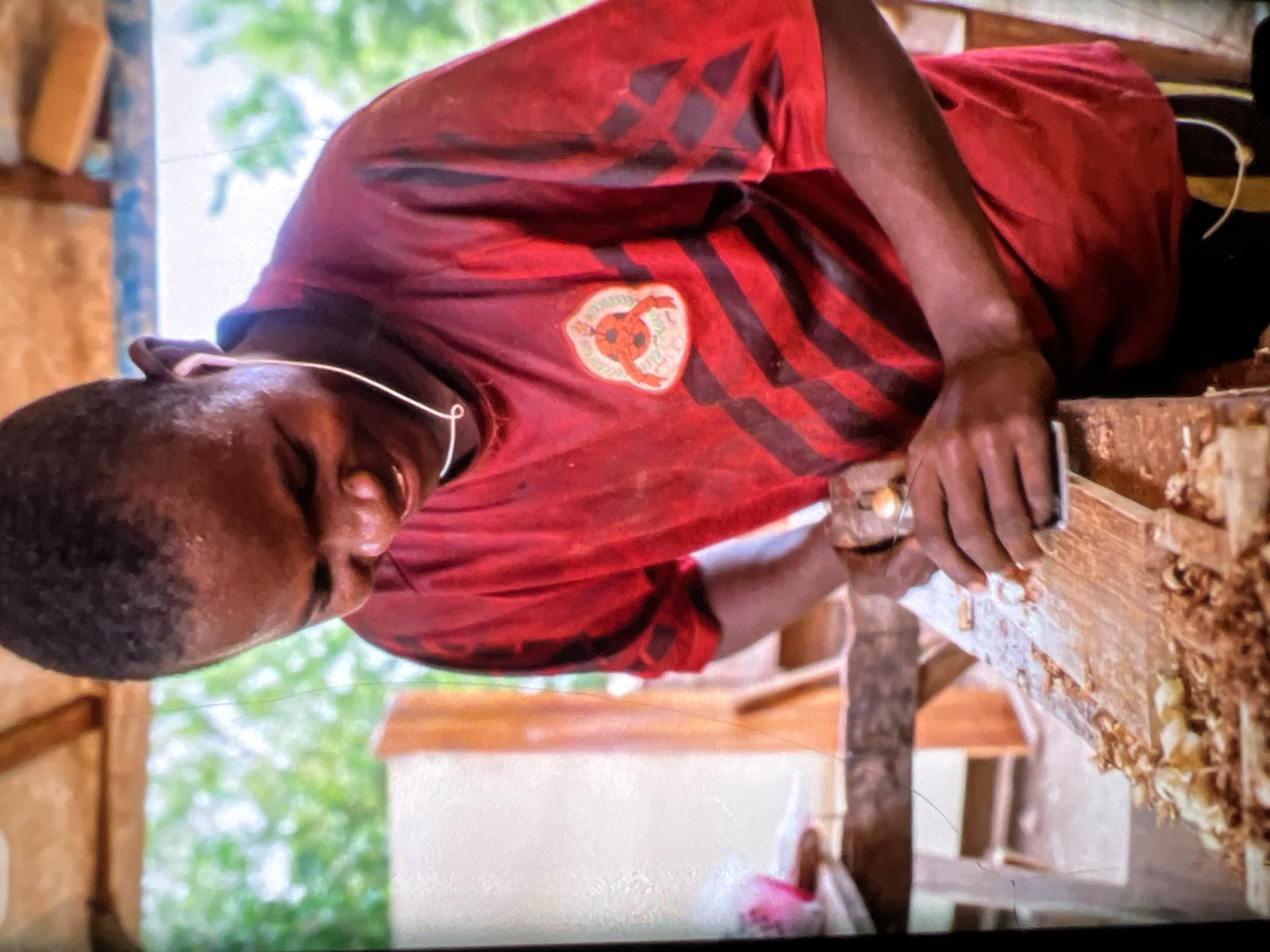 A young Black male, wearing a red t-shirt with black stripes and a circular emblem on the left chest, is seen from the chest up, working with a woodworking plane. He is smiling and looking down at a wooden board covered in wood shavings. He has white earbuds in his ears. The background consists of wooden beams and greenery, suggesting an outdoor or semi-outdoor workshop setting. The scene appears to be in Lagos, Nigeria, given the implied context of the location. The lighting is natural, casting soft shadows.