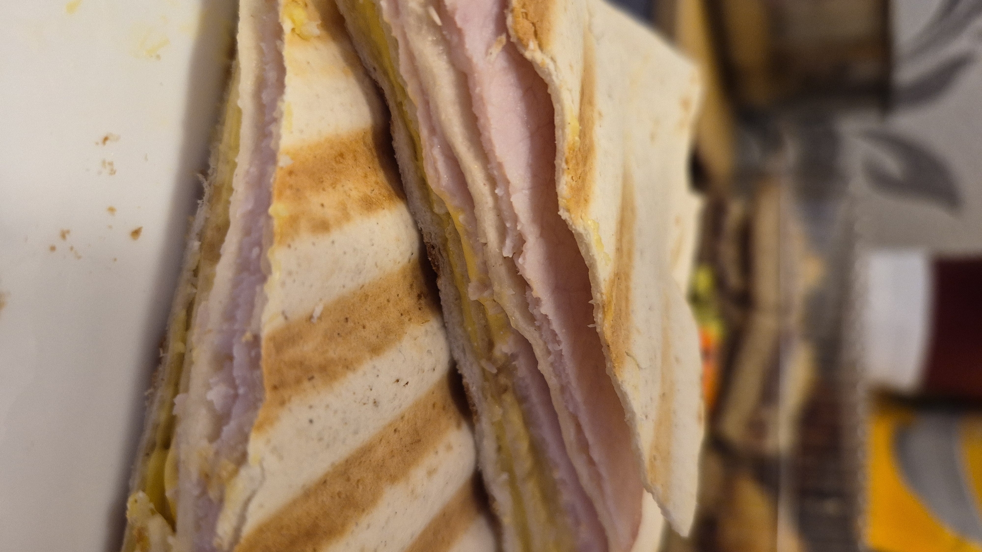 The image shows a close-up, eye-level view of a grilled sandwich, likely a panini or quesadilla, resting on a white surface. The sandwich is cut in half, revealing layers of thin-sliced pink ham and a yellow filling, possibly cheese or mustard. The bread is golden brown with distinct grill marks. The background is blurred, suggesting it is out of focus, and shows what appears to be kitchen appliances and containers. The lighting is dim, with warm tones. There is no visible indication of the city of Krauja, Latvia, nor any people or actions occurring. The scene is static and focused on the food item.