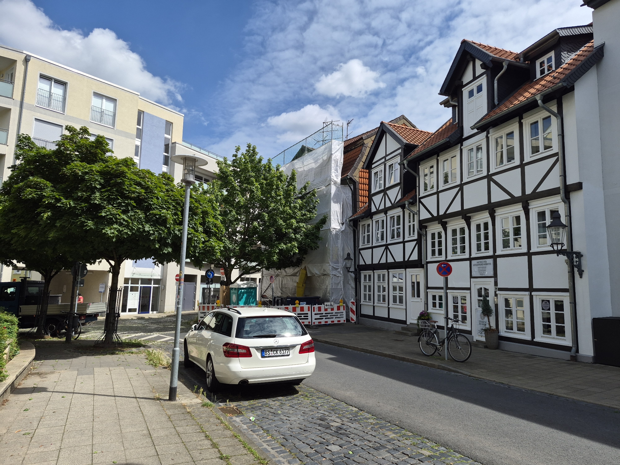 The image depicts a street scene in Brunswick, Germany, specifically showing a section of the city that features both modern and historic architecture. On the left side of the street, a modern apartment building with a pale yellow and blue facade stands tall. In front of this building, several large green trees provide shade. Adjacent to the modern building is a street with a paved sidewalk.
The right side of the street showcases traditional German half-timbered houses with white plaster walls and dark wooden beams. These houses appear to be occupied by businesses, as indicated by signage visible on some of the buildings. One building displays a sign that reads "Praxis für Medicinische Kosmetik" (Practice for Medical Cosmetics).
A white station wagon is parked on the side of the road. A bicycle is leaning against the facade of one of the half-timbered houses. Further down the street, scaffolding and white sheeting suggest that a building is undergoing renovation or construction. The sky is blue with scattered white clouds, indicating a pleasant day. The street itself is paved with cobblestones in some areas and asphalt in others. Traffic signs and street lamps are also present.