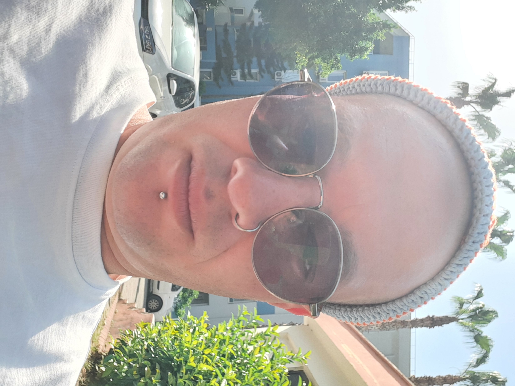 A man is wearing sunglasses and a headband in Alanya, Turkey. He is wearing a white t-shirt and has a piercing on his lip. Behind him is a building, a white car, green foliage, and palm trees. The image is oriented at an angle, suggesting it was taken as a selfie. The bright sunlight suggests a daytime setting.