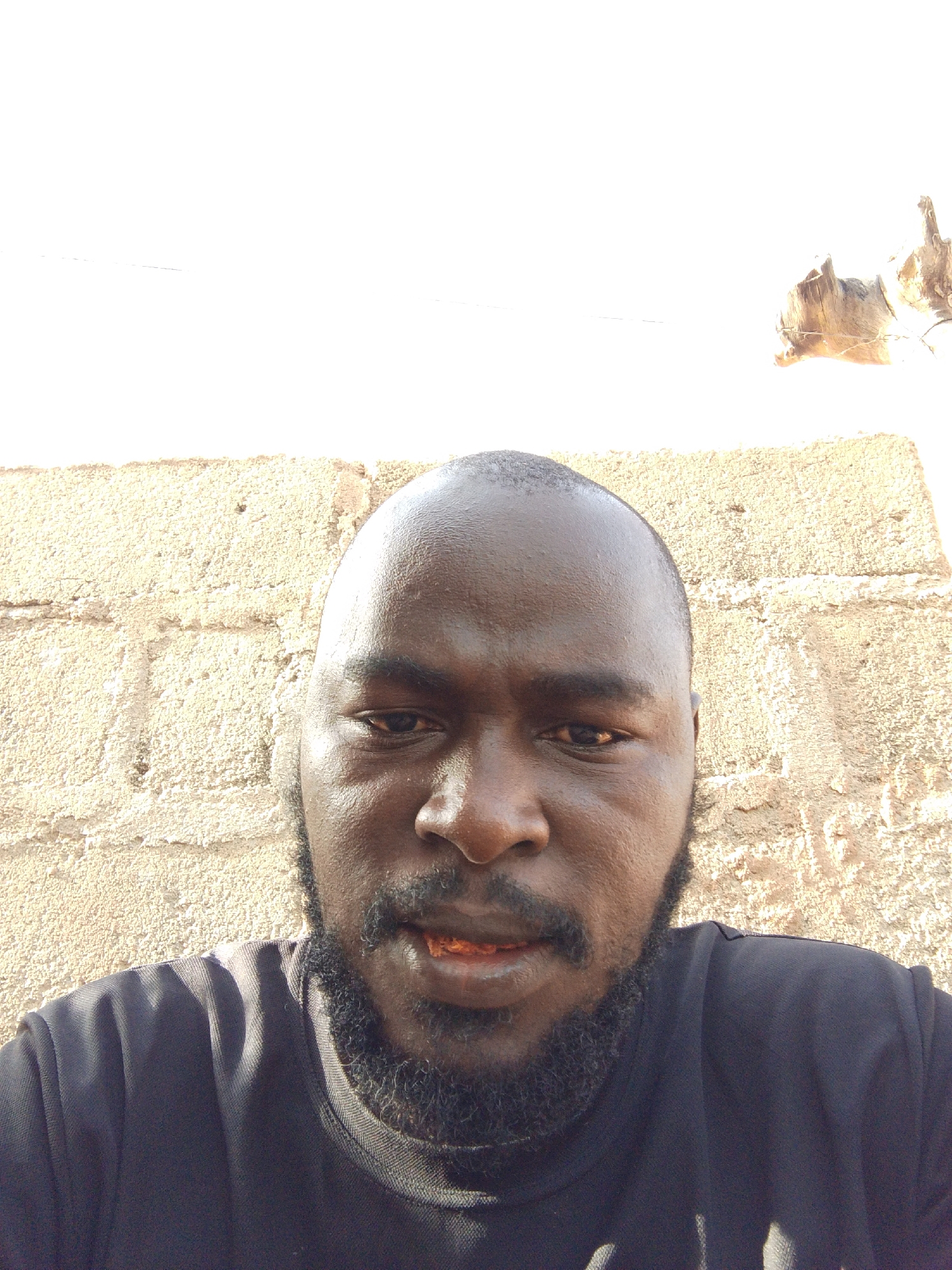 A close-up, eye-level shot depicts a dark-skinned man with a shaved head and a full, dark beard, positioned in front of a light-colored, textured wall. The man is wearing a dark t-shirt. A small, reddish-orange object is visible in his mouth. Based on the location information, the photo was taken in Mulke Barikin Sayejo, Nigeria.