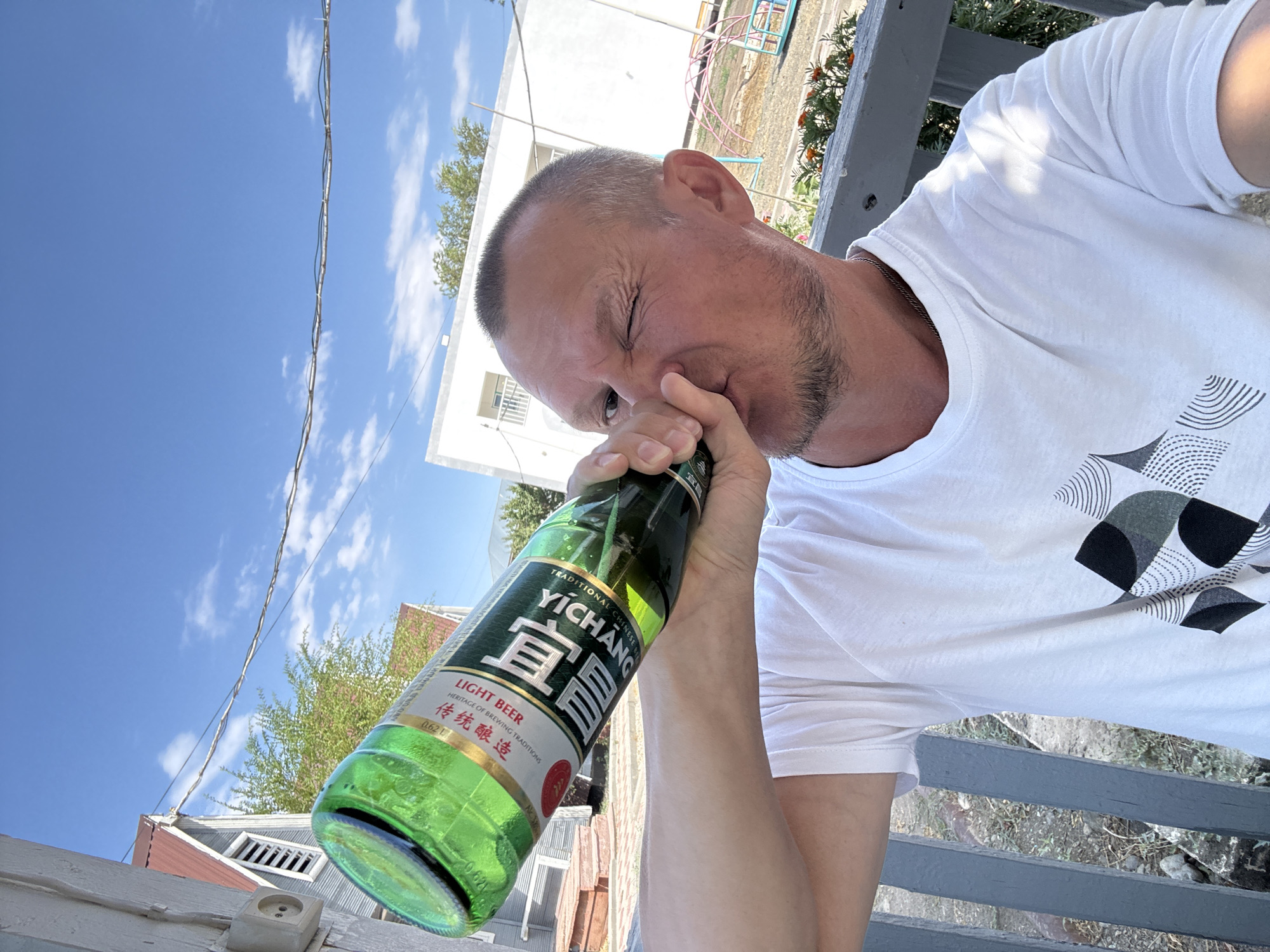 A fair-skinned man with short-cropped hair and a light beard is drinking from a green glass bottle of Yichang Traditional Chinese Beer. He is wearing a white t-shirt with a black and grey geometric pattern on the chest. He is outdoors, seated on a gray wooden bench, with a blurred background showing a white building with a balcony, a red-roofed structure, trees, and a bright blue sky with scattered clouds. A power outlet is visible on the white wooden siding of a building to the left. The image appears to be taken on a sunny day. The specific location is Zharbulak, Kazakhstan.