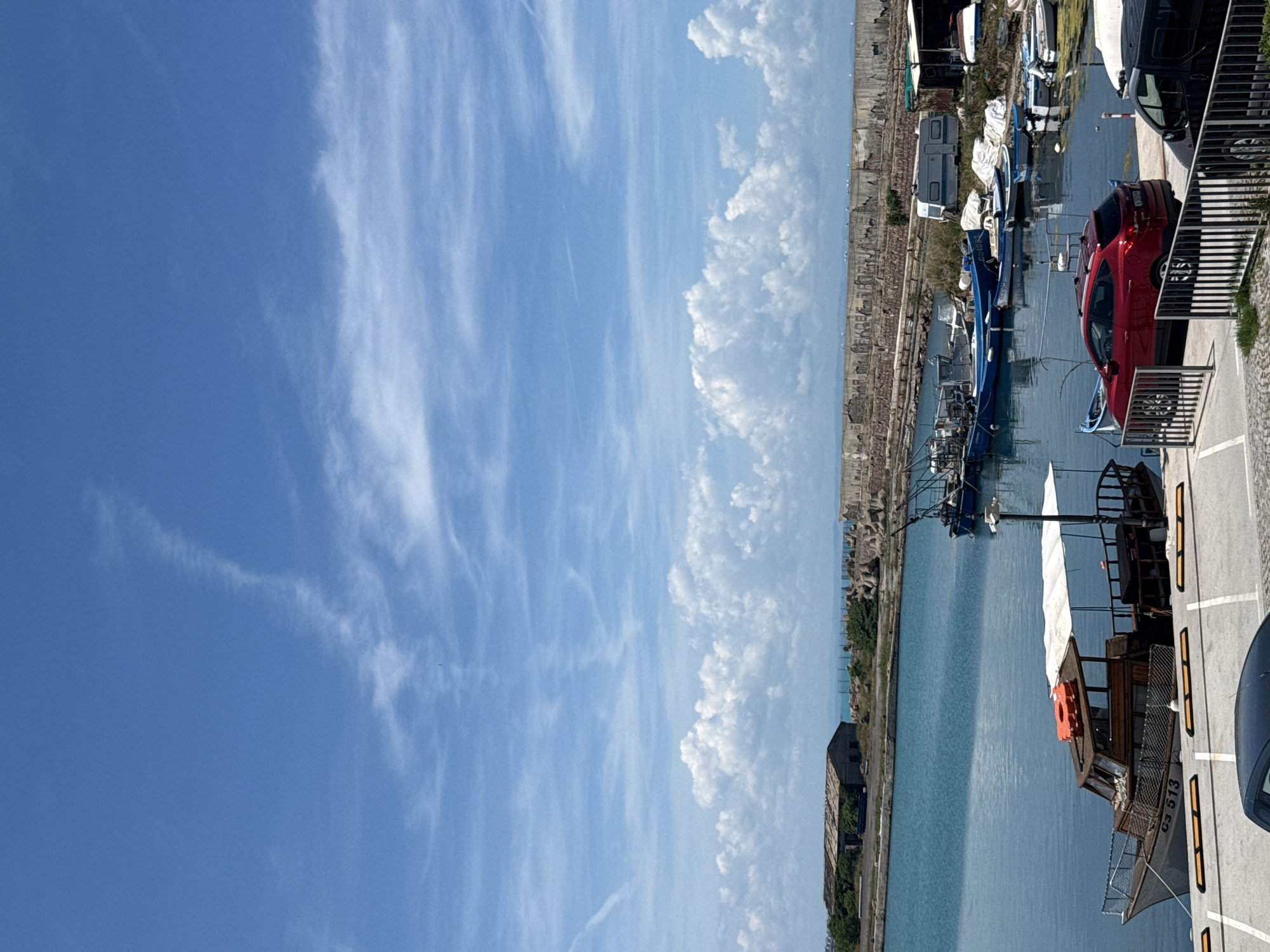 A wide-angle vertical shot captures a scenic view of the harbor in Sozopol, Bulgaria on a bright, sunny day. The clear blue sky is adorned with scattered white clouds, some of which are puffy cumulus formations. Below the sky, a vast expanse of tranquil blue water fills the lower half of the image, reflecting the sky and the surrounding elements.
Several boats are docked in the harbor. In the foreground, a wooden boat with a white canopy and a life preserver on its roof is prominently featured, displaying the registration number "G3 513". To its right, a red SUV is parked, and behind it, a black vehicle is partially visible. Further out in the water, a collection of smaller blue and white fishing boats are moored. In the background, a long, weathered stone breakwater stretches across the horizon. On the land side of the harbor, various structures are visible, including a dilapidated building with a corrugated roof, a white caravan, and some makeshift shelters. The overall scene depicts a typical day at a bustling fishing harbor.