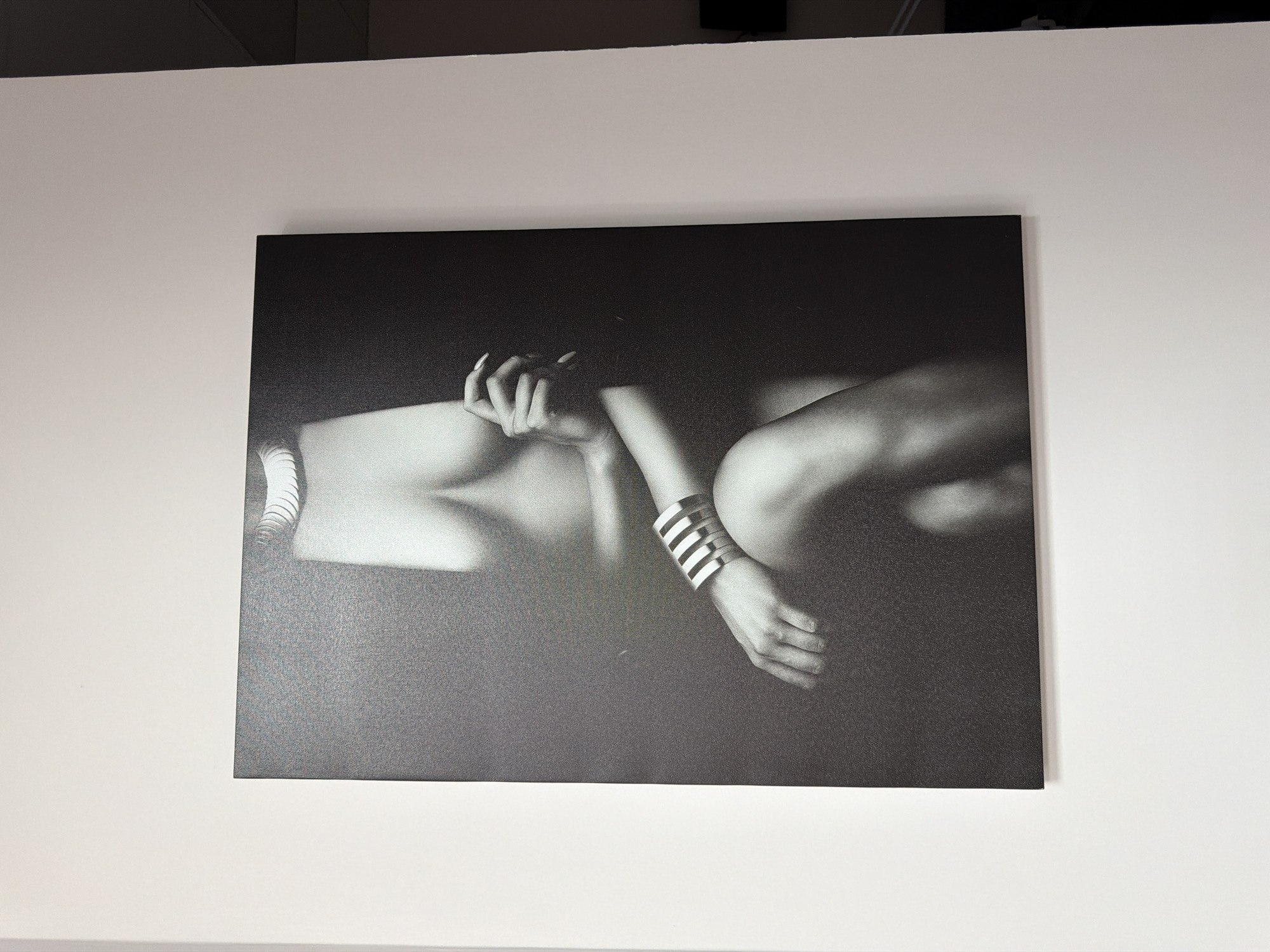 A black and white photograph hangs on a white wall. The photograph depicts a nude figure, possibly female, with a wrist adorned with a cuff-style bracelet composed of horizontal bars. The figure's form is softly lit, creating shadows and highlights that emphasize curves. The background appears dark and indistinct. The setting is an interior space. The location is Unknown, Unknown.