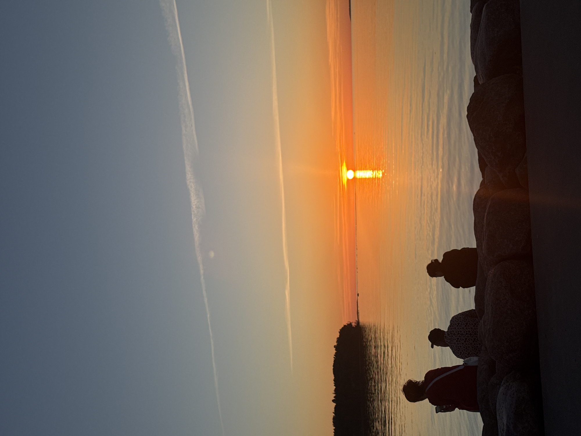 This image captures a sunset over a body of water in Tallinn, Estonia. The sky gradient transitions from a deep twilight blue at the top to vibrant orange and yellow at the horizon. Two distinct white contrails from aircraft are visible; one extends diagonally across the mid-upper sky, and another shorter, more diffused one is higher and to the left. The sun, a bright white-yellow disc, is positioned low on the horizon, slightly right of the frame's center, creating a prominent vertical reflection path on the calm water. A dark, tree-covered landmass is silhouetted against the sunset on the left side of the water, with a faint, darker landmass visible on the far right horizon. In the immediate foreground, three individuals are seated on a line of large, dark rocks, presented in silhouette with their backs partially or fully to the viewer, facing the sunset. The leftmost person wears a red garment, a shoulder bag, and holds a white cup. The central individual wears a patterned shirt and a backward baseball cap. The rightmost person, appearing smaller, has dark clothing and their hair tied up. Below the rocks, a dark, flat paved surface is visible.