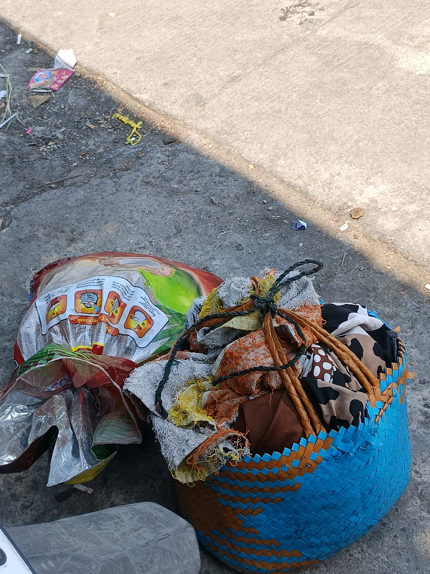 In Blega, Indonesia, a close-up shot reveals a blue and orange woven basket filled with various fabric items, including patterned cloth and frayed material. A string ties the fabrics together. Next to the basket lies a large, crinkled plastic bag, partially opened, with colorful graphics and instructional images. The items are situated on a concrete surface, which appears to be a sidewalk or paved area, with patches of dirt and scattered debris. A shadow cuts across the scene diagonally from the upper right, suggesting it is daytime. In the background, a portion of a motorcycle or similar vehicle is visible in the lower left corner.
