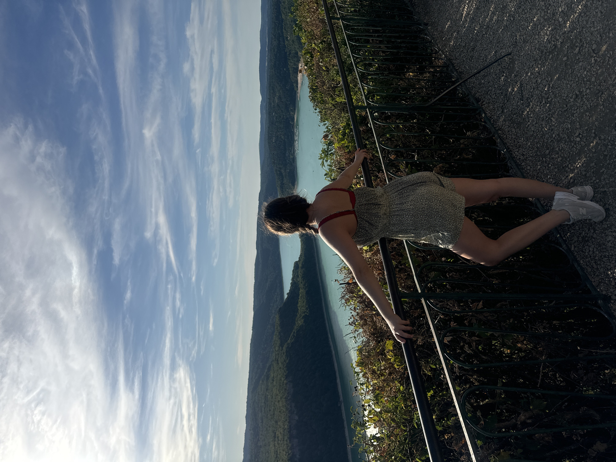 A woman with braided dark hair stands on a gravel walkway, leaning on a black metal railing. She wears a light-colored floral romper and grey sneakers, facing away from the camera towards a vast, turquoise body of water bordered by lush green hills. The sky above is a clear blue with wispy white clouds. The scene suggests a scenic overlook, likely in the vicinity of Moirans-en-Montagne, France, given the characteristic landscape of the Jura region.