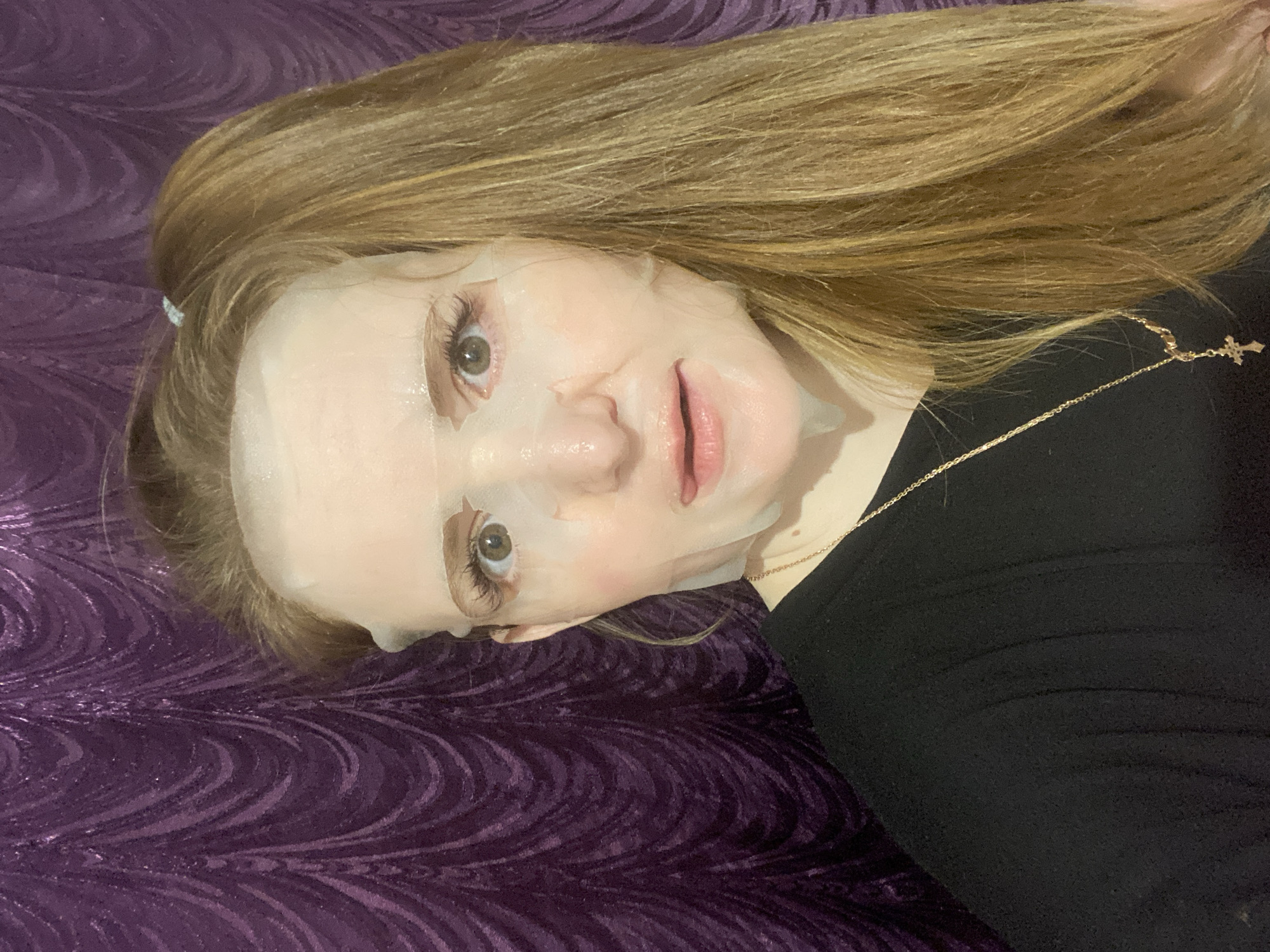 A light-skinned person with reddish-blonde hair is lying on a textured purple surface. They are wearing a black shirt and a gold necklace with a cross pendant. A translucent facial sheet mask covers their face. The location is unknown, and there's no indication it's in Chaikovsky, Russia.
