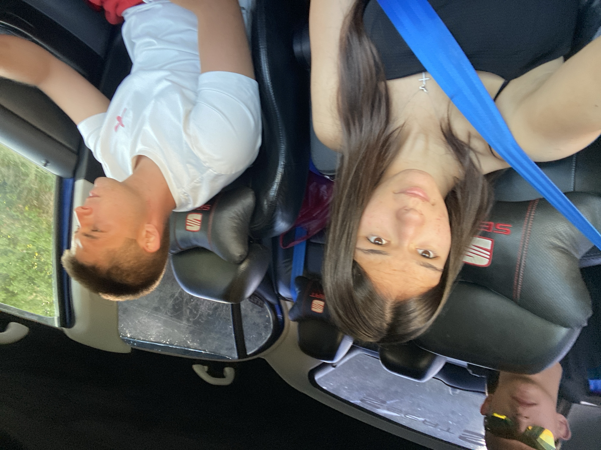 Four people are inside a car. In the front passenger seat, a young woman with long brown hair and a black top is wearing a silver cross necklace and a bright blue seatbelt. To her right, a young man in a white t-shirt is driving. In the back seat, a person with short blonde hair wearing sunglasses is visible, and another person's head is partially visible behind the driver. The car seats have "SEAT" branding on them, suggesting it is a SEAT vehicle. The interior of the car is dark, with light filtering in from the windows, revealing green foliage outside. The scene appears to be a road trip on a sunny day. The question implies the location is Sevlievo, Bulgaria, but this information is not visually determinable from the image.