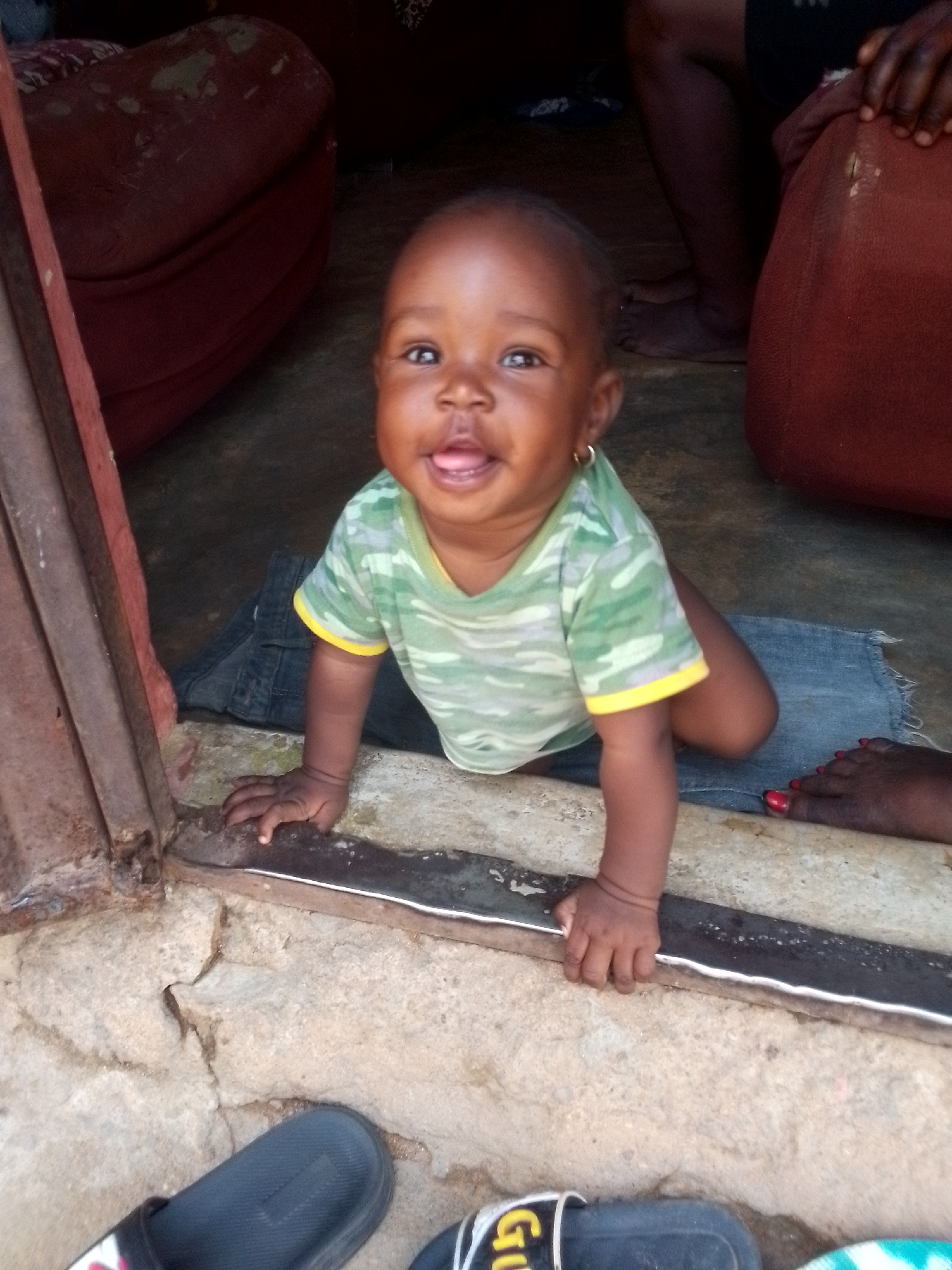 A baby wearing a green and yellow camouflage-patterned shirt is crawling towards the camera. The baby is on a blue denim cloth. The baby has one gold hoop earring in one ear and has their tongue slightly sticking out. The baby is on the floor in front of a door with a brown frame. Outside the door, there is cracked concrete and two slides. Red furniture can be seen inside the room. Another person is nearby, with red nail polish visible on one foot. The location is likely Narayi, Nigeria.