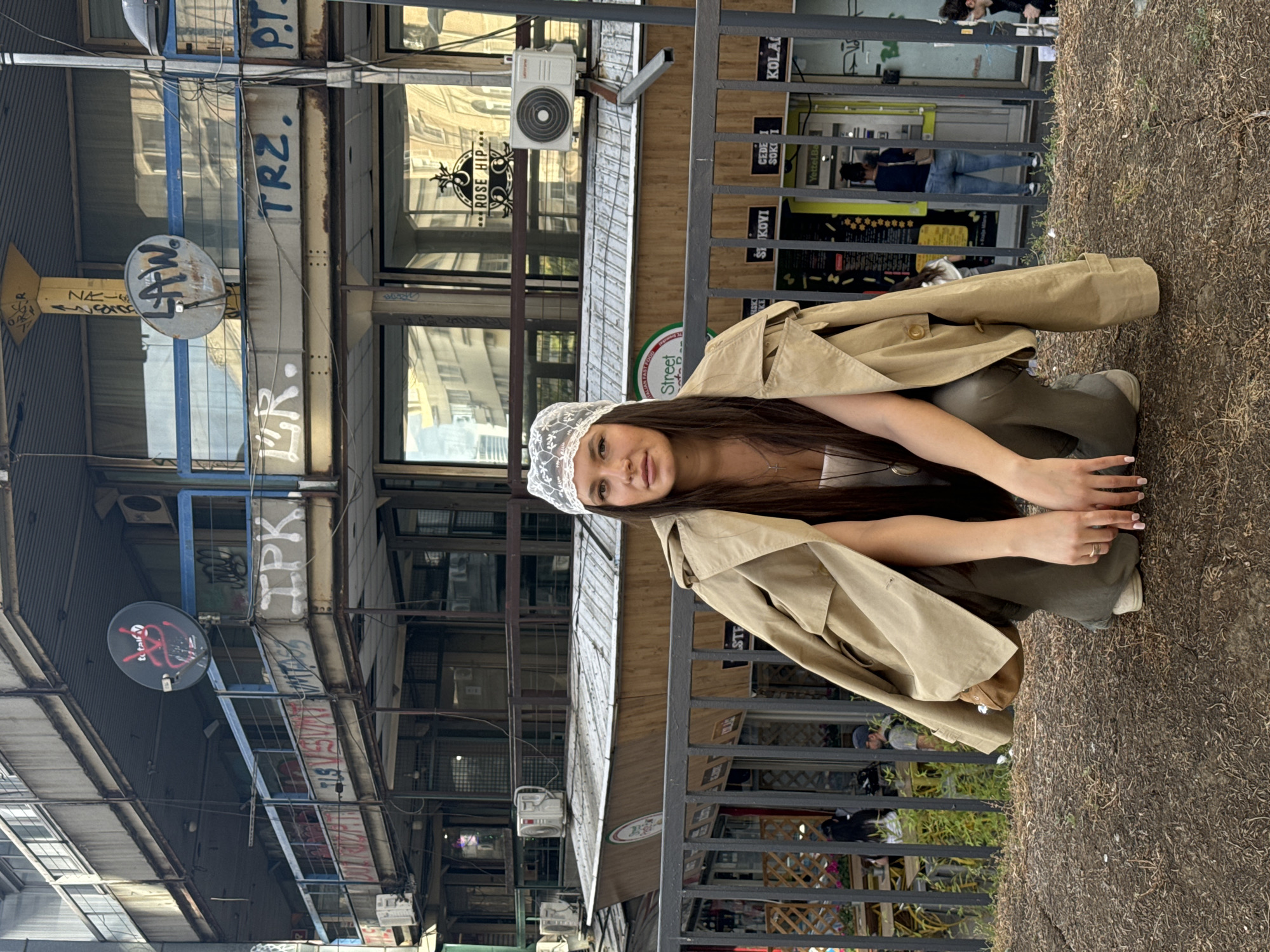 The image depicts a young woman crouching behind a railing outdoors. She is wearing a lacy white cap, a neutral-toned top, olive green pants, and a tan trench coat draped over her shoulders. She is looking at the camera with her hands placed in front of her.
In the background, there is a densely packed urban environment. Buildings with visible signage in Serbian are present, some of which include "KOLAC", "CEDE I SOKI", "SI KOVI", and "Yettel.Ba". Graffiti and satellite dishes are affixed to the buildings. The signage “ROSE HIP” is also visible. The street has power lines and other urban infrastructure overhead.
The setting appears to be an alleyway or a narrow street. Pedestrians can be seen walking in the background. The environment is indicative of Belgrade, Serbia.