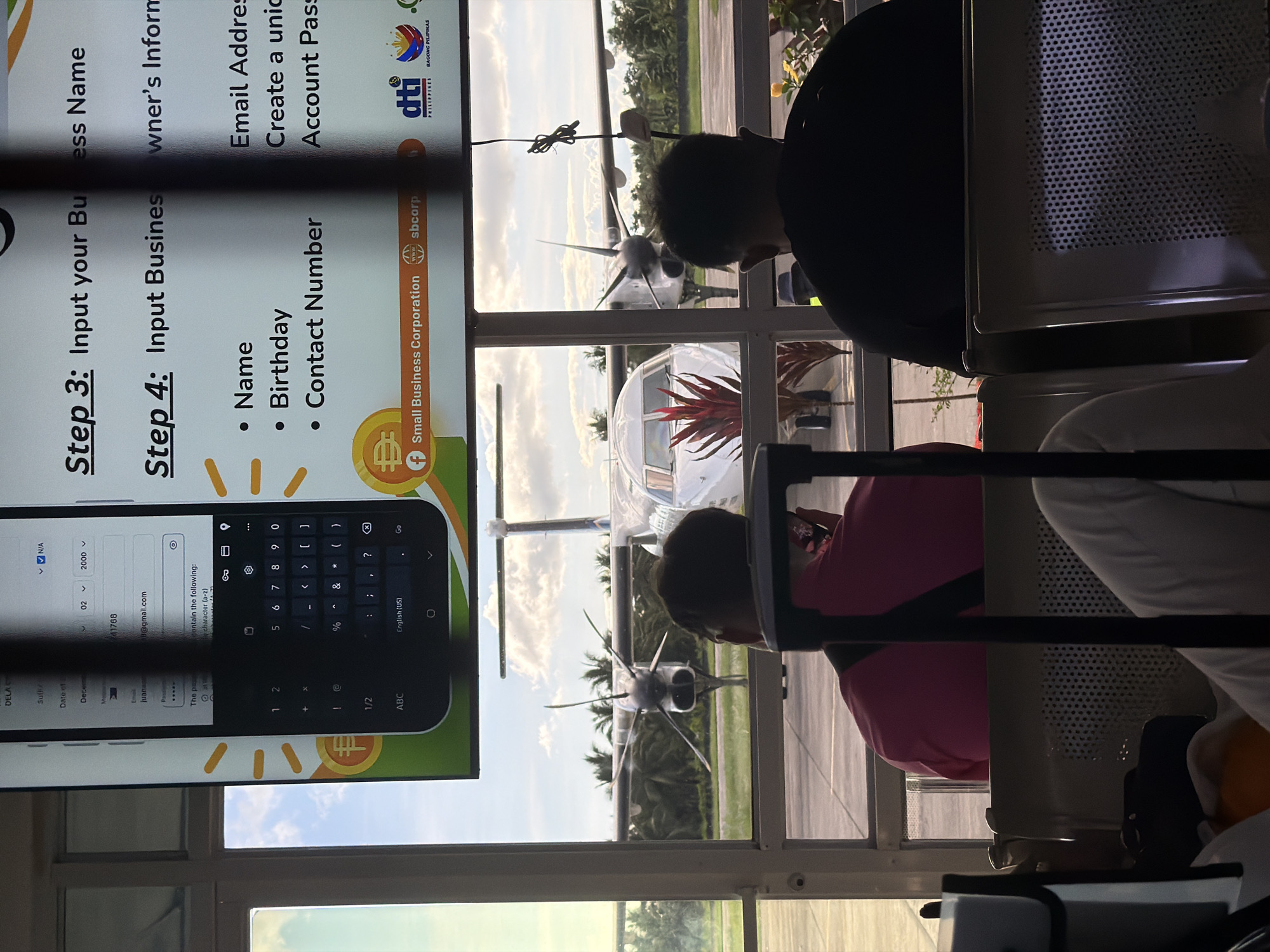 The image shows an airport waiting area, possibly in Del Carmen, Philippines, based on the DTI logo displayed on a screen. Visible through the window is a small aircraft on the tarmac. Inside, people are sitting on waiting chairs, partially obscuring the view of the airplane. A screen displays a digital form, with prompts for inputting business information such as business name, owner's information, email address, name, birthday, and contact number. A digital keyboard is visible on the screen.