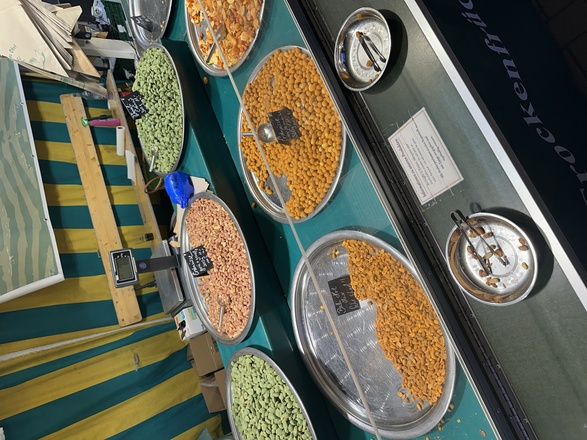The image shows a market stall in Bruchhausen-Vilsen, Germany, featuring various types of nuts displayed in large metal trays. The stall is set up with a green and yellow striped awning in the background, and a wooden bench behind the trays. There is a digital scale on the bench, along with some boxes and cleaning supplies.
In the foreground, there are several trays of nuts. The leftmost tray contains green nuts, possibly wasabi peas. Next to it is a tray of pinkish-brown nuts, identified by a sign as "Erdnüsse geröstet" (roasted peanuts). To the right of the peanuts is a tray of bright orange nuts, with a sign that reads "Bali Hot Mix" and "Sehr scharf!!" (very spicy!!). Another tray, further to the right, contains a mix of nuts and other items. In the bottom right of the image, a small metal bowl with tongs holds a few scattered nuts.
A sign placed on the counter provides information about the products, including a note about allergens and the use of tongs for hygiene. The overall scene depicts a typical food market stall offering a selection of seasoned and roasted nuts.
