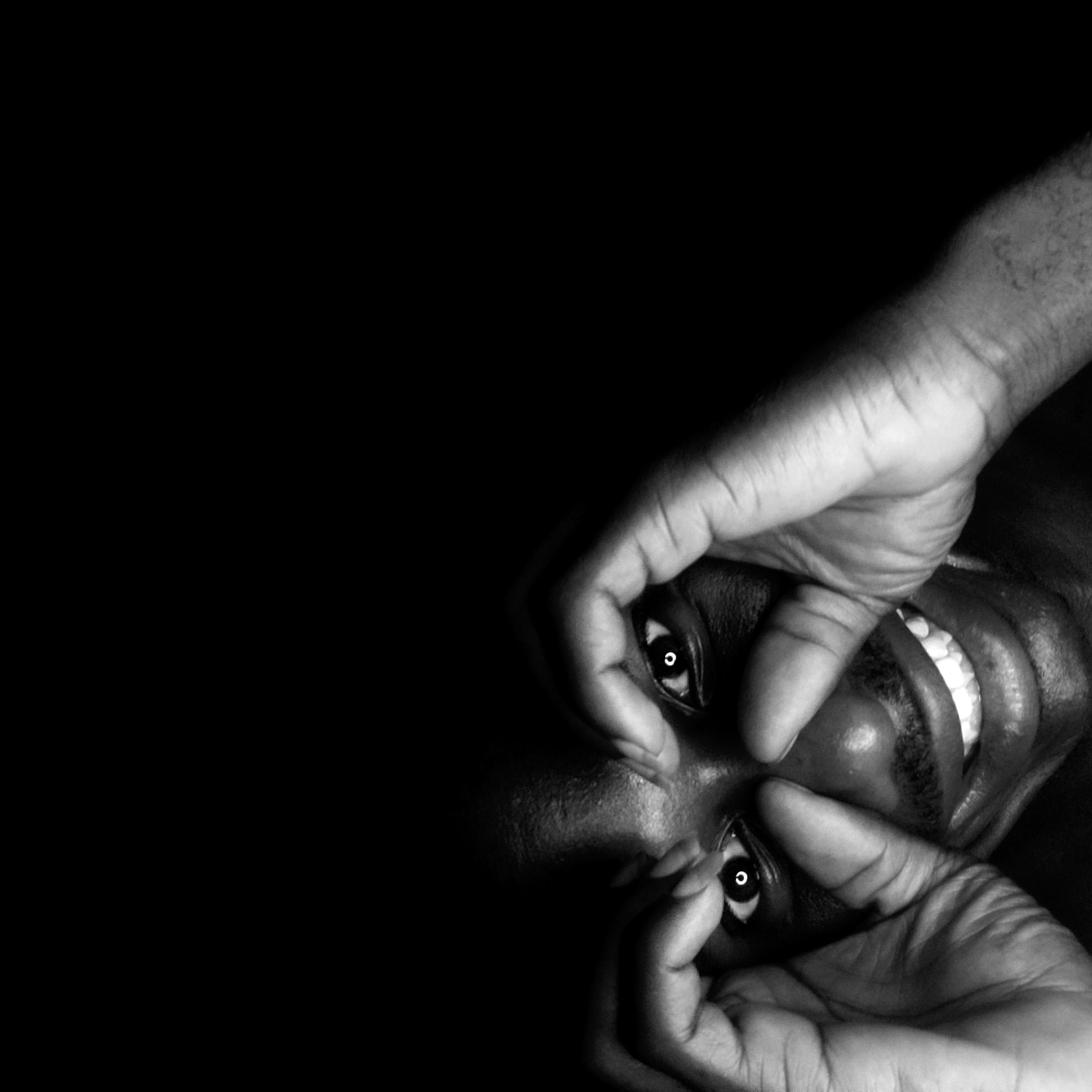 A black and white photograph depicts a person's face with two hands forming circles around the eyes, creating a makeshift pair of spectacles. The person is smiling, revealing their teeth. The background is dark. The image does not provide enough details to determine the location as Narayi, Nigeria.