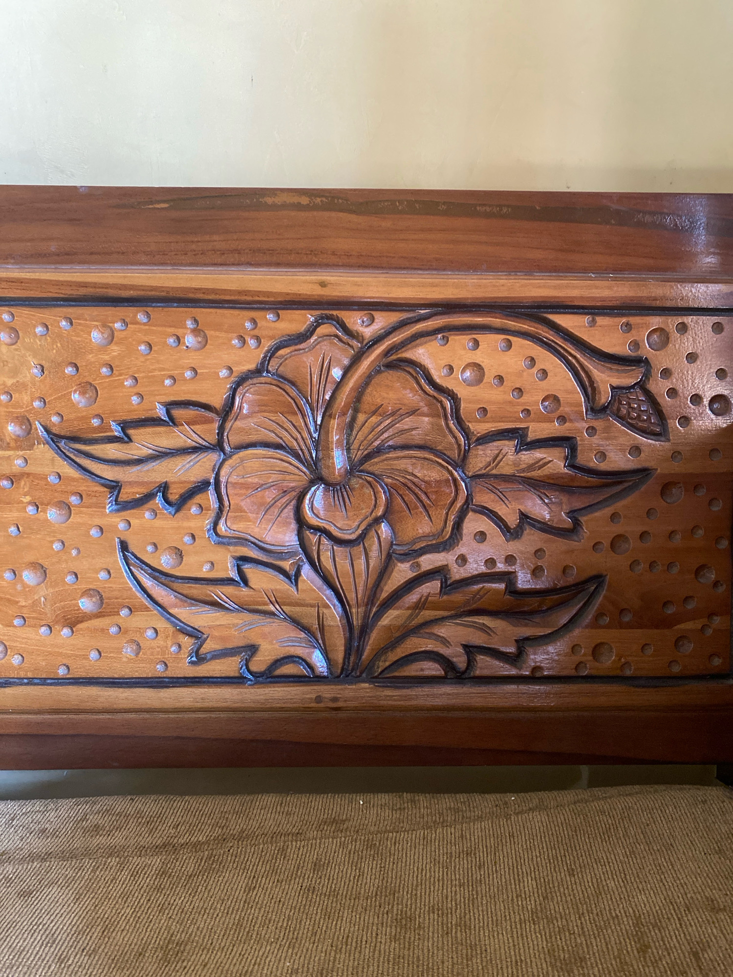 This is a close-up, eye-level, medium shot of a carved wooden panel on a piece of furniture, likely a headboard or a decorative panel. The carving depicts a prominent, stylized flower with several large petals and intricate details, surrounded by smaller, rounded, raised circular shapes that resemble bubbles or dew drops. The wood appears to be a dark, rich brown with a glossy finish, and the carved details are highlighted with darker shading. The panel is framed by darker wood trim. The background consists of a pale yellow wall above the wooden furniture and a textured, light brown woven fabric below. The scene is indoors, suggesting it is a domestic or hospitality setting. There are no people or animals visible in the image. The overall impression is one of craftsmanship and decorative artistry. The location in Jimbaran Kulon, Indonesia, suggests a likely Balinese influence in the carving style, which is common in this region for wood crafts.
