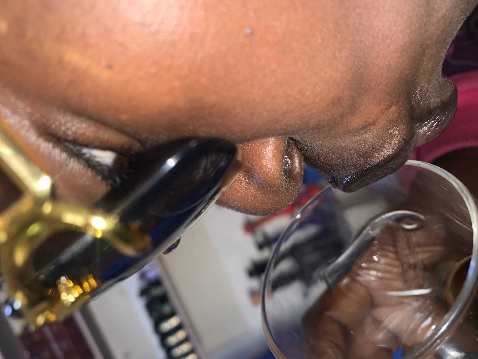 The image shows a close-up, angled shot of a person's mouth, nose, and chin, with what appears to be a straw and a glass in the foreground. The person has dark skin and appears to be wearing sunglasses with gold frames. The background is blurred, but suggests an indoor setting with some shelves and bottles. The lighting is warm and seems to be artificial, possibly from indoor lights or a flash. The overall mood is casual. There is no visible text in the image.