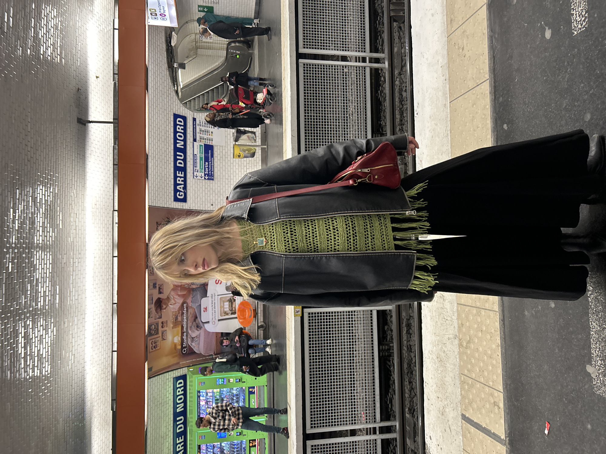 The image depicts a woman standing on a platform at Gare du Nord, a major train station in Paris, France. She is wearing a black jacket over a green crocheted top with fringe, a long black skirt, and boots. A red bag is slung over her shoulder. Behind her, the station's signage is visible, reading "GARE DU NORD" and directing passengers to services and exits. Other people are visible in the background, walking and waiting. The environment includes train tracks, a platform edge, and tiled walls, with advertisements and vending machines in view.