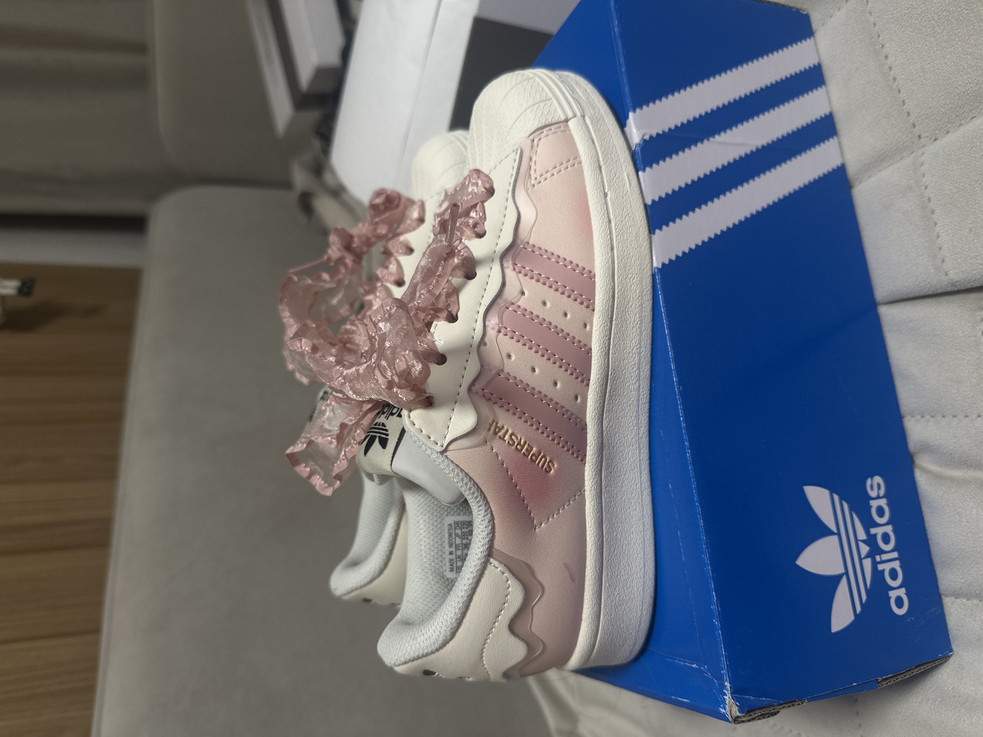 The image shows a pair of pink and white Adidas Superstar sneakers with ruffled, pink laces resting on top of a blue Adidas shoebox. The shoebox is placed on a light-colored, textured surface, possibly a couch or bed. The image is taken indoors. There is no evidence to suggest the location is in Votkinsk, Russia, or any city for that matter. The photo focuses on the shoes and box.