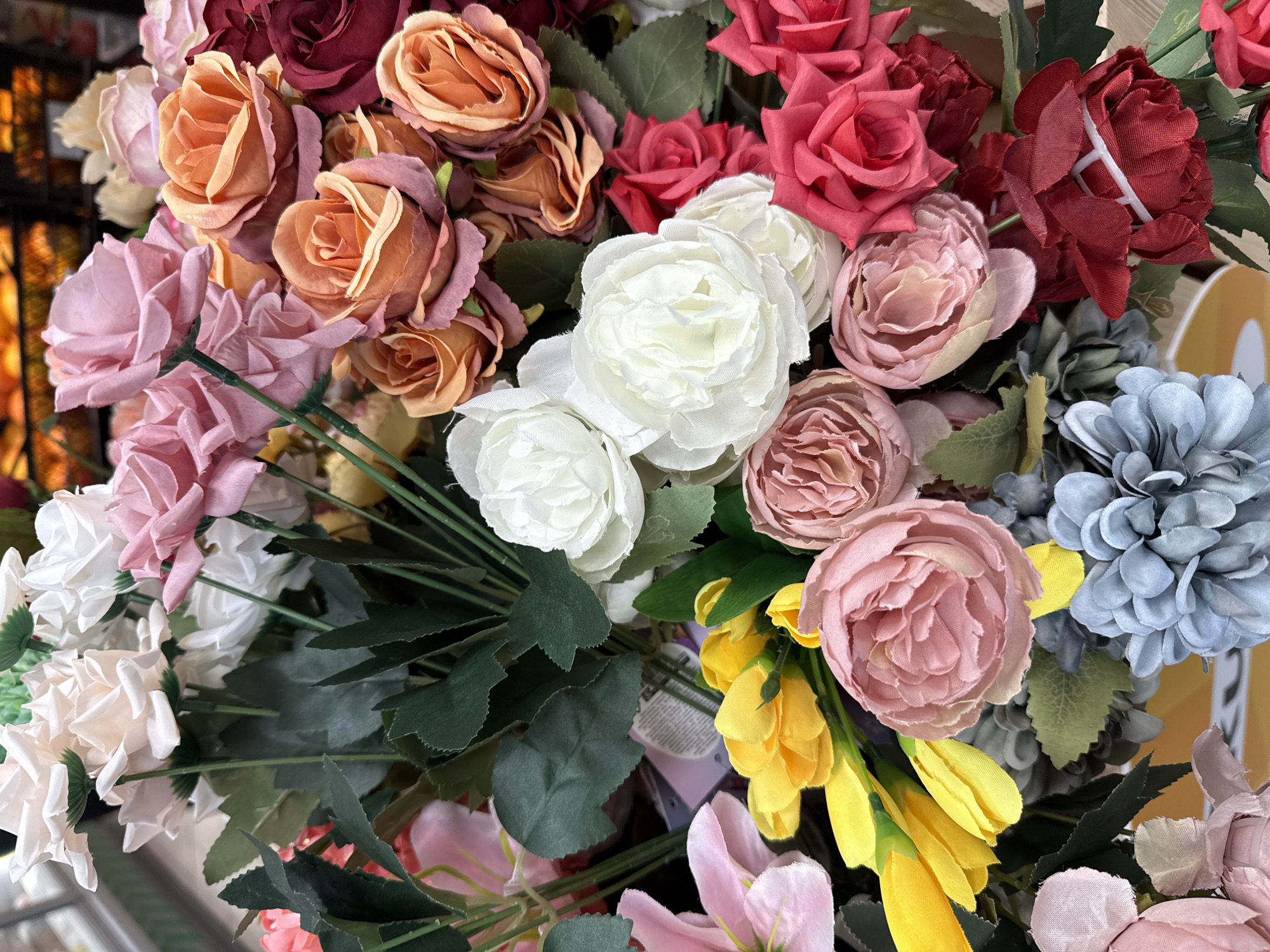 The image shows a close-up view of a bouquet of artificial flowers. The bouquet contains various colors and types of flowers, including roses (red, pink, white, peach), blue hydrangeas, and yellow tulips. There are also green leaves interspersed among the flowers. The overall composition fills the frame.
The scene does not contain any indications of location in Votkinsk, Russia, or any indication of human presence or interaction.