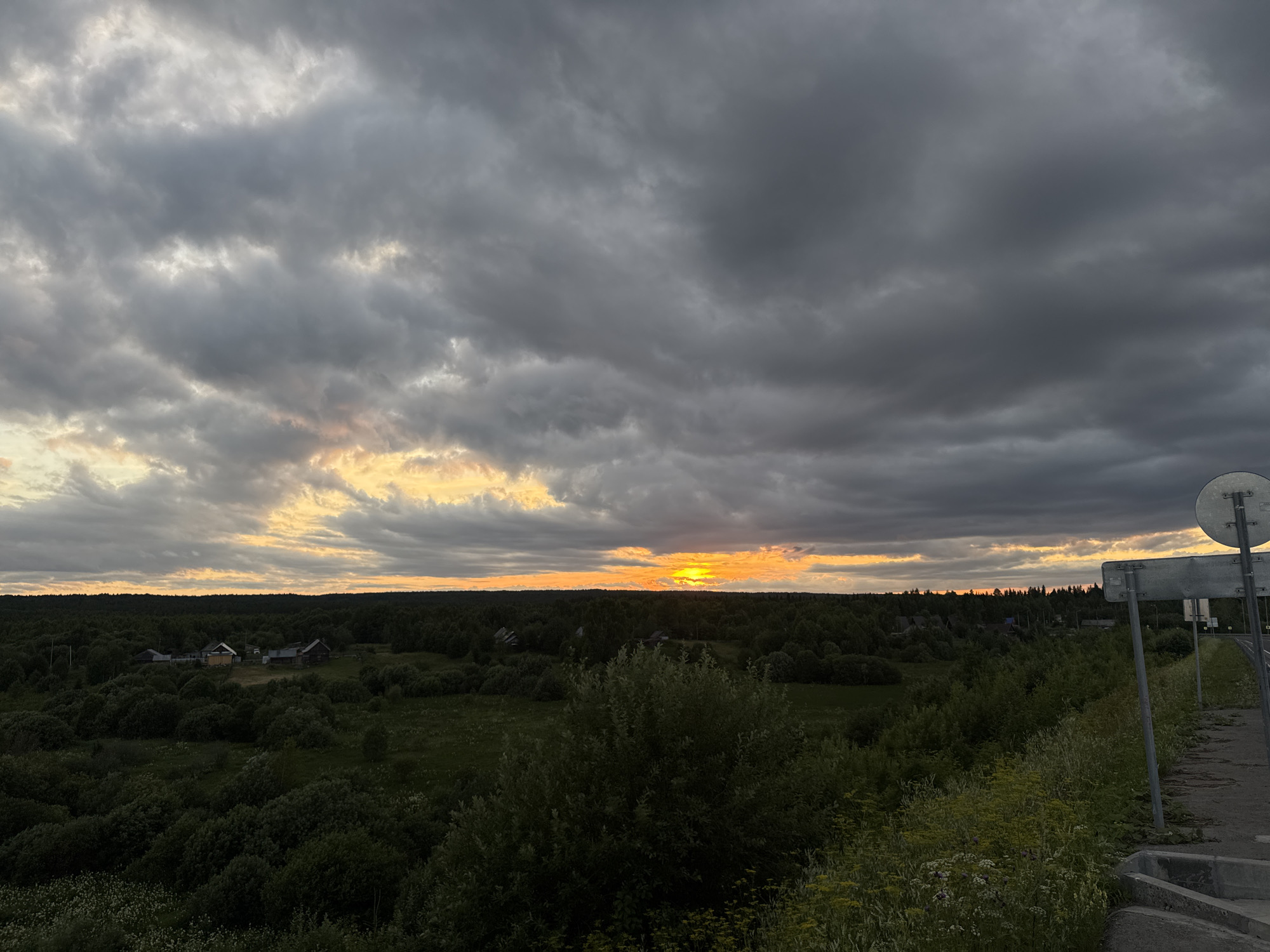 The image shows a wide landscape view from a slightly elevated position, looking out over a rural area in Ryabinino, Russia, during sunset. The sky is dominated by heavy, dark grey clouds with patches of warm orange and yellow light breaking through near the horizon, indicating the setting sun. A line of dense, dark green trees stretches across the middle ground, obscuring much of the landscape beyond. Within the tree line, a small cluster of houses with visible roofs can be seen. In the foreground, there is a patch of green vegetation and wildflowers, with a paved surface and some signage on the right side of the frame, suggesting a roadside or overlook. The overall atmosphere is dramatic due to the contrasting light and dark elements in the sky and landscape.