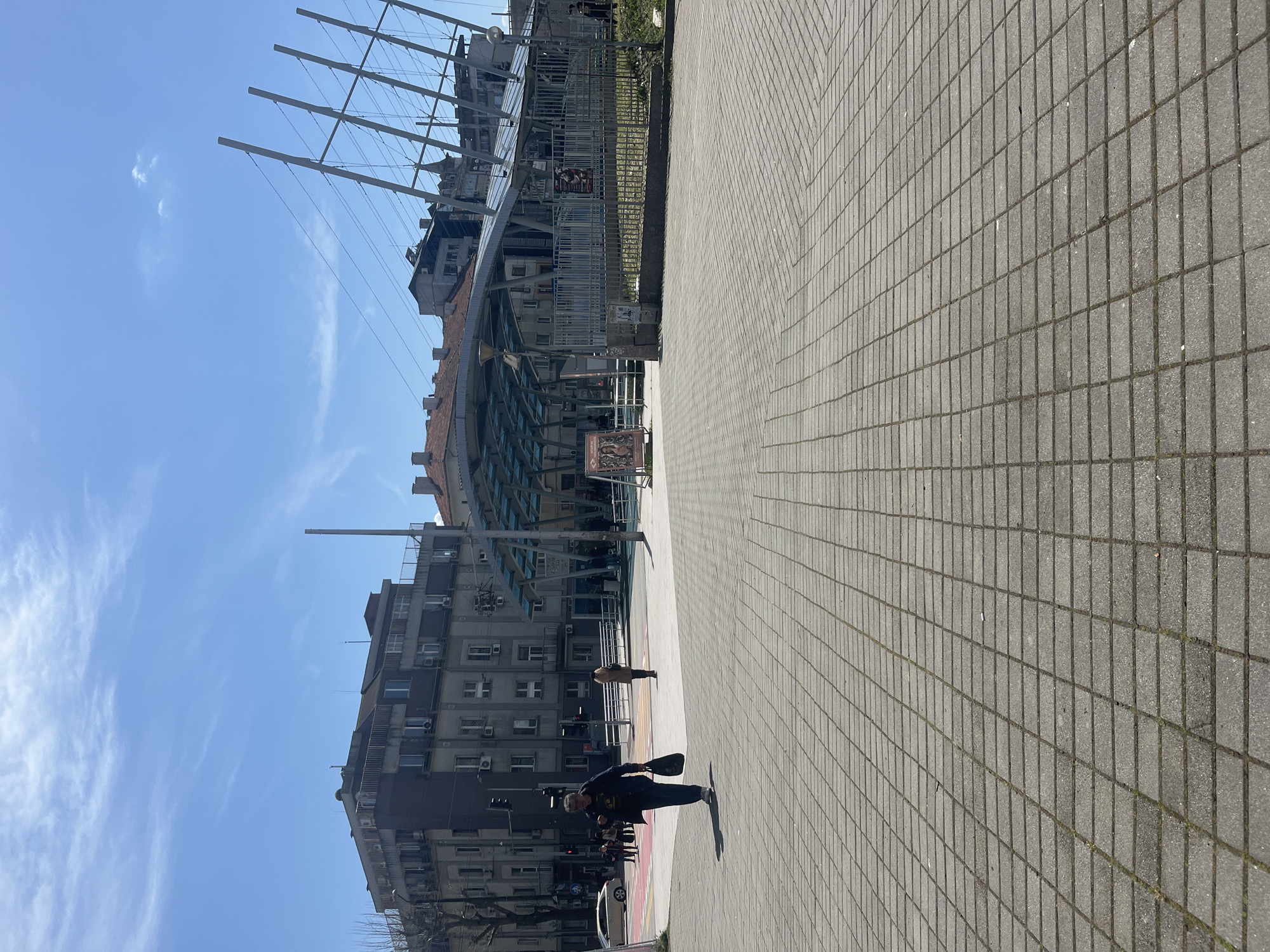The image shows a street scene in Burgas, Bulgaria, featuring a paved walkway with a pedestrian walking across it. In the background, there is an old warship on display, the "Nadiejda" (Hope) frigate of the navy of Bulgaria. Buildings are visible on the left side of the frame, under a clear blue sky.
