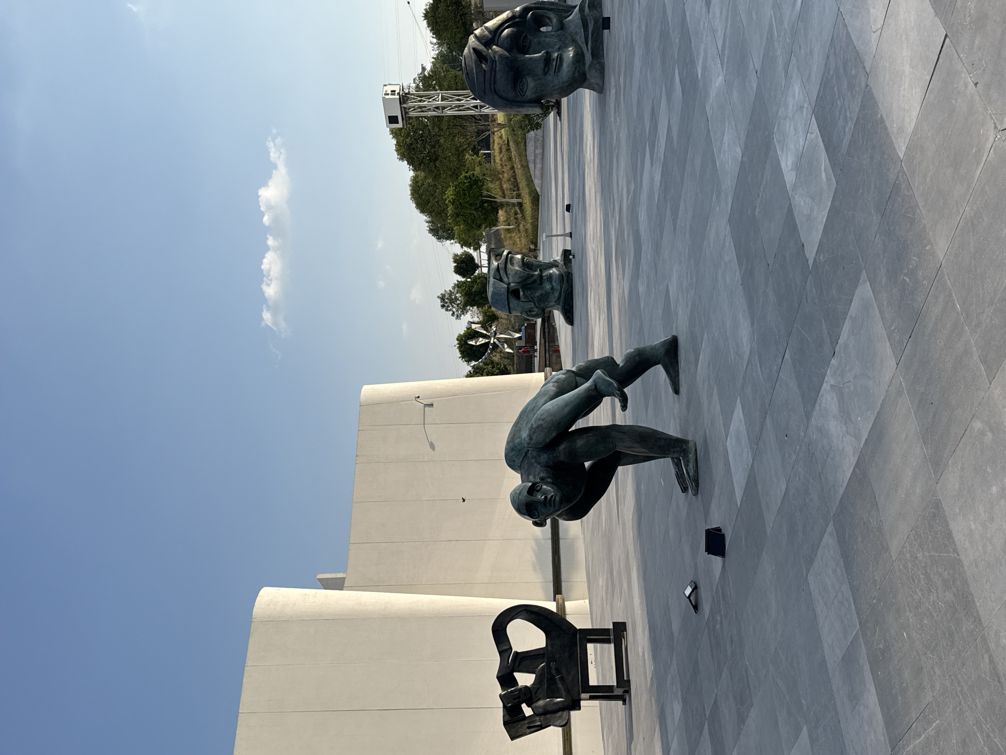 The image shows several bronze statues on a paved surface against a building and a blue sky in San Bernardino Tlaxcalancingo, Mexico. There are three sculptures depicting human heads and a sculpture of two figures in a difficult human balancing act, in which one person is supporting the other. In addition, there is a small cubist bronze on a pedestal. In the background, a tall, gray tower rises above the tree line.