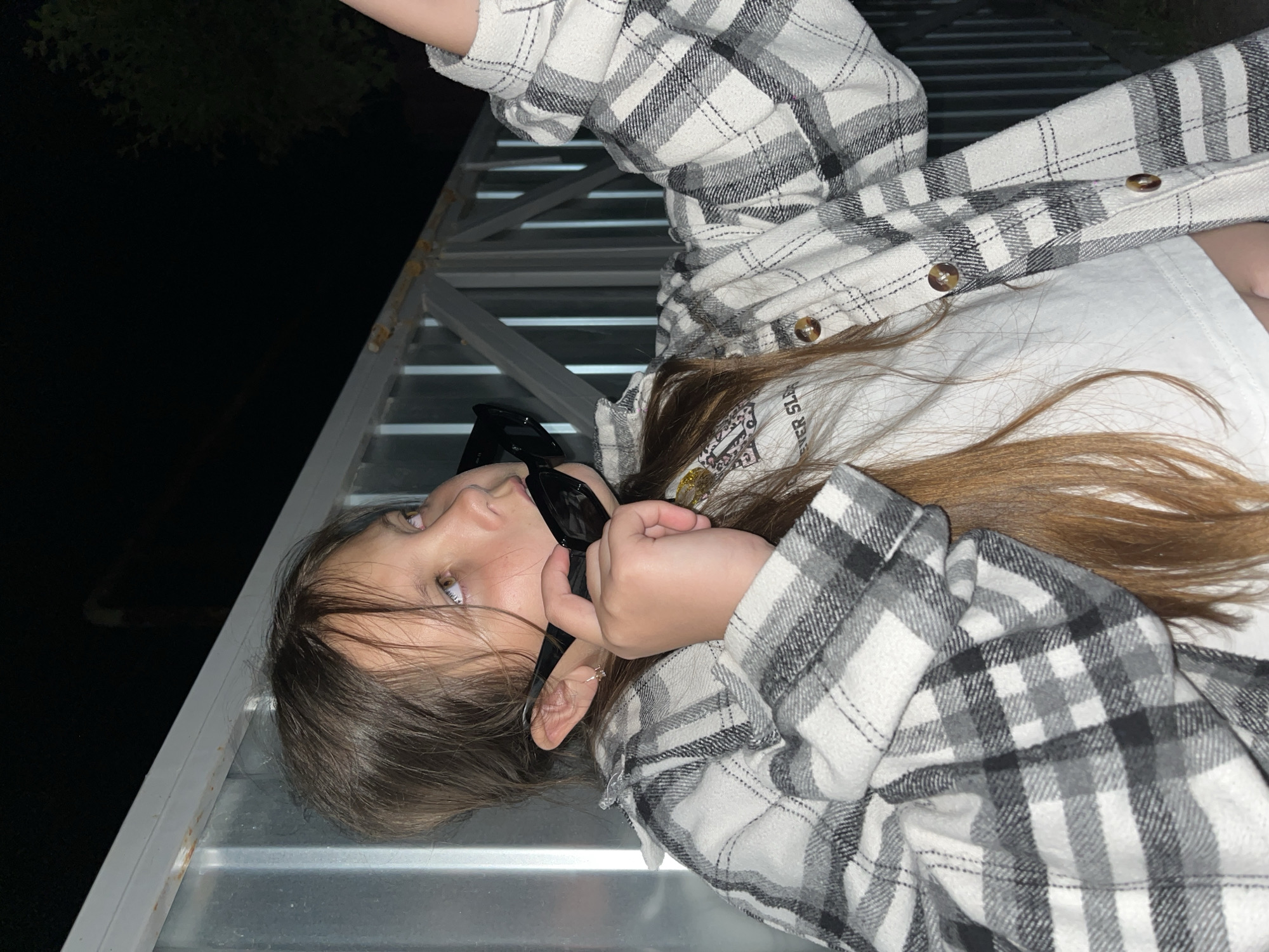 A young person is lying on a metal structure at night. The person is wearing a plaid shirt over a white t-shirt and holding sunglasses to their face. The background is dark, possibly a night scene in Novopokrovskaya, Russia, though there is no specific information from the image to confirm this.