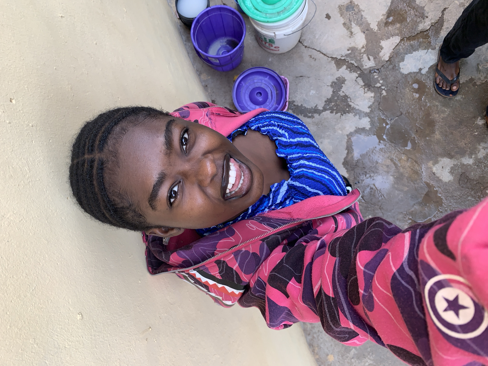 A young Black woman smiles directly at the camera, appearing to take a selfie. She is wearing a pink and purple patterned hooded jacket and a blue striped shirt. The background includes a light beige wall to her left and a concrete area to her right. Several buckets of different colors are visible behind her. A person wearing black pants and sandals stands near the right edge of the image. The location is Rafin Par, Nigeria.