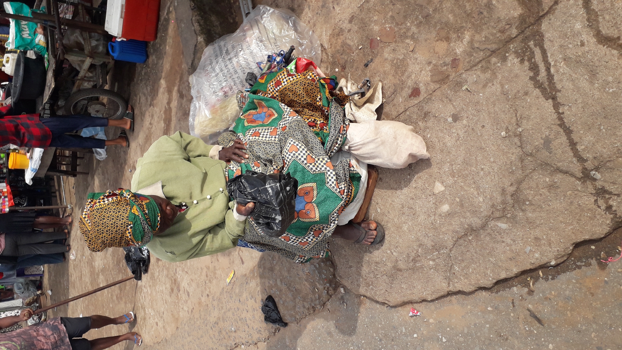 This image shows an overhead view of a person sitting on the ground, surrounded by various objects. The person is wearing a green long-sleeved shirt and a headwrap with a yellow and black patterned fabric. They are also covered by a colorful, patterned cloth, predominantly green with black and orange motifs. A black plastic bag is held in their hands. They are wearing sandals.
The scene appears to be a market or roadside setting in Nkpor, Nigeria. Scattered around the person are piles of fabric, a large clear plastic bag filled with what looks like food or other goods, and a white sack. In the background, a cart with a wheel is visible, along with various market stalls and people walking. The ground is concrete and somewhat weathered, with cracks and some damp areas.
The lighting suggests it is daytime, likely with bright sunlight given the shadows. The overall atmosphere seems to be one of everyday life, possibly a vendor resting or waiting for customers. There is no clear indication of specific emotions, but the scene depicts a person in a public space surrounded by their belongings. No legible text is visible in the image.