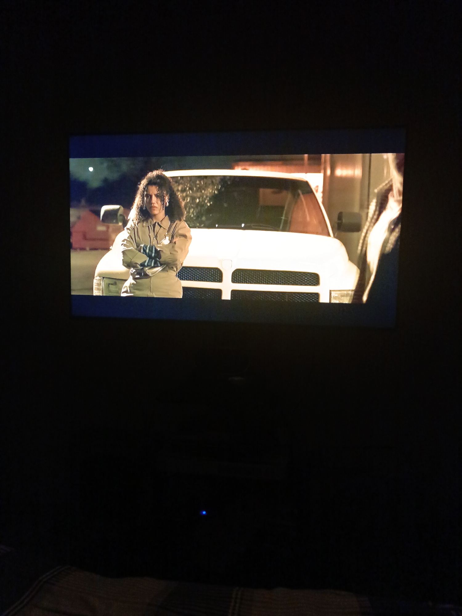 The media file shows a still from a television screen. In the center of the screen, a woman with curly dark hair stands with her arms crossed, looking directly at the camera with a serious expression. She is wearing a tan jumpsuit and appears to be outdoors at night, with a white vehicle behind her. The vehicle is a truck or van, and its headlights are on, illuminating the area. The background is dark and out of focus, but hints of buildings or structures can be seen. The overall lighting suggests a nighttime setting, possibly in an urban or industrial area. The image is grainy and has a slightly bluish tint, indicating it is a capture of a screen rather than a direct photograph. There is a small blue light visible in the dark area below the screen, possibly from a device.