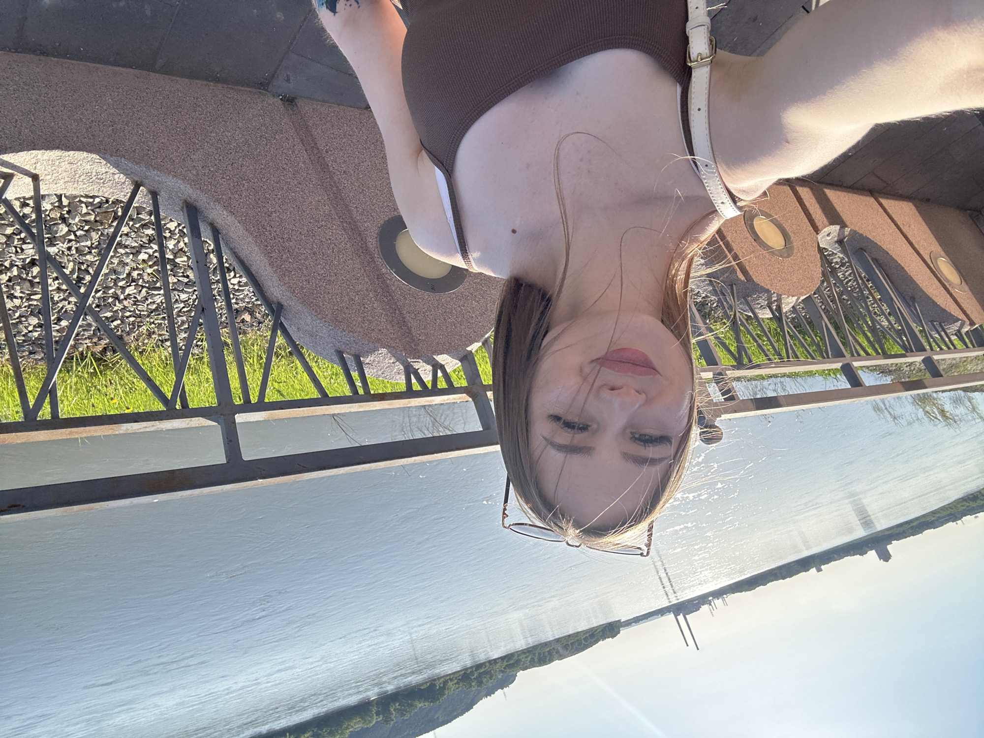 A young woman is captured in a selfie taken from above, looking down towards the camera with a neutral expression. Her light brown hair is slightly windswept, and she is wearing a dark brown ribbed tank top with a white bra strap visible. A white shoulder bag strap is also visible over her right shoulder. She has glasses resting on her face. A tattoo is visible on her upper left arm.
She is standing on a textured, light brown paved surface, likely a promenade or embankment, which features recessed circular lights. A dark metal railing separates this area from a wide body of water, appearing to be a river or large lake. Below the railing, grass and gravel are visible.
The background is dominated by the expansive water, which reflects strong sunlight. On the distant horizon, a strip of land with trees and some tall, slender structures (possibly industrial elements or bridge components) can be seen. The sky is bright and clear, indicating a sunny day. There is no visible text in the image.