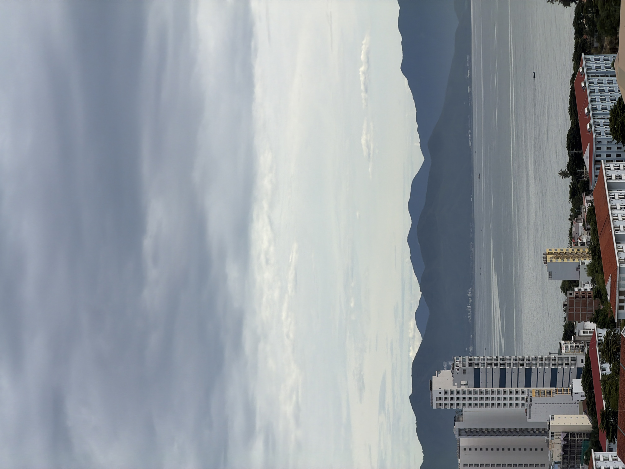The image captures a cloudy day over the city of Nha Trang, Vietnam, with a view of the coast. In the foreground, several multi-story buildings are visible, some with modern architectural designs and others appearing older. One prominent white high-rise building stands out, characterized by its numerous windows and blue horizontal stripes. To the left, another tall white building is partially visible.
The middle ground is dominated by a body of water, likely the sea, stretching towards a backdrop of silhouetted mountains. Several small boats are scattered across the water's surface. The mountains themselves are layered, with darker peaks in the distance fading into a hazy sky.
The sky above is filled with a blanket of overcast clouds, ranging in tone from light gray to darker, more dense patches, suggesting an impending change in weather or recent rainfall. The overall lighting is diffused, characteristic of an overcast day, casting a subdued light on the urban landscape and the natural elements. There are no people clearly visible in the image.