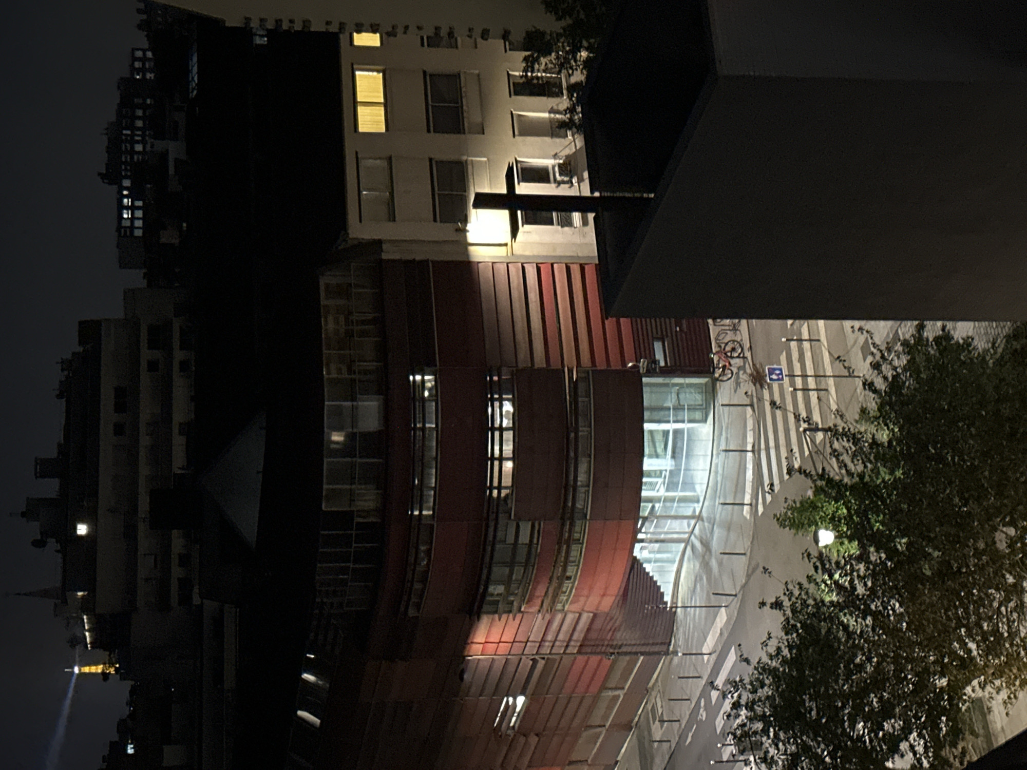 The image shows a nighttime scene in Paris, France. The Eiffel Tower is visible in the distance. In the foreground, there are several buildings, including a modern structure with curved, red and metallic facade. A white building with visible windows is situated on top of the red structure. A street with a bicycle is visible to the right of the building, along with a tree. A street lamp illuminates the foliage. A large, dark architectural element looms above the view.