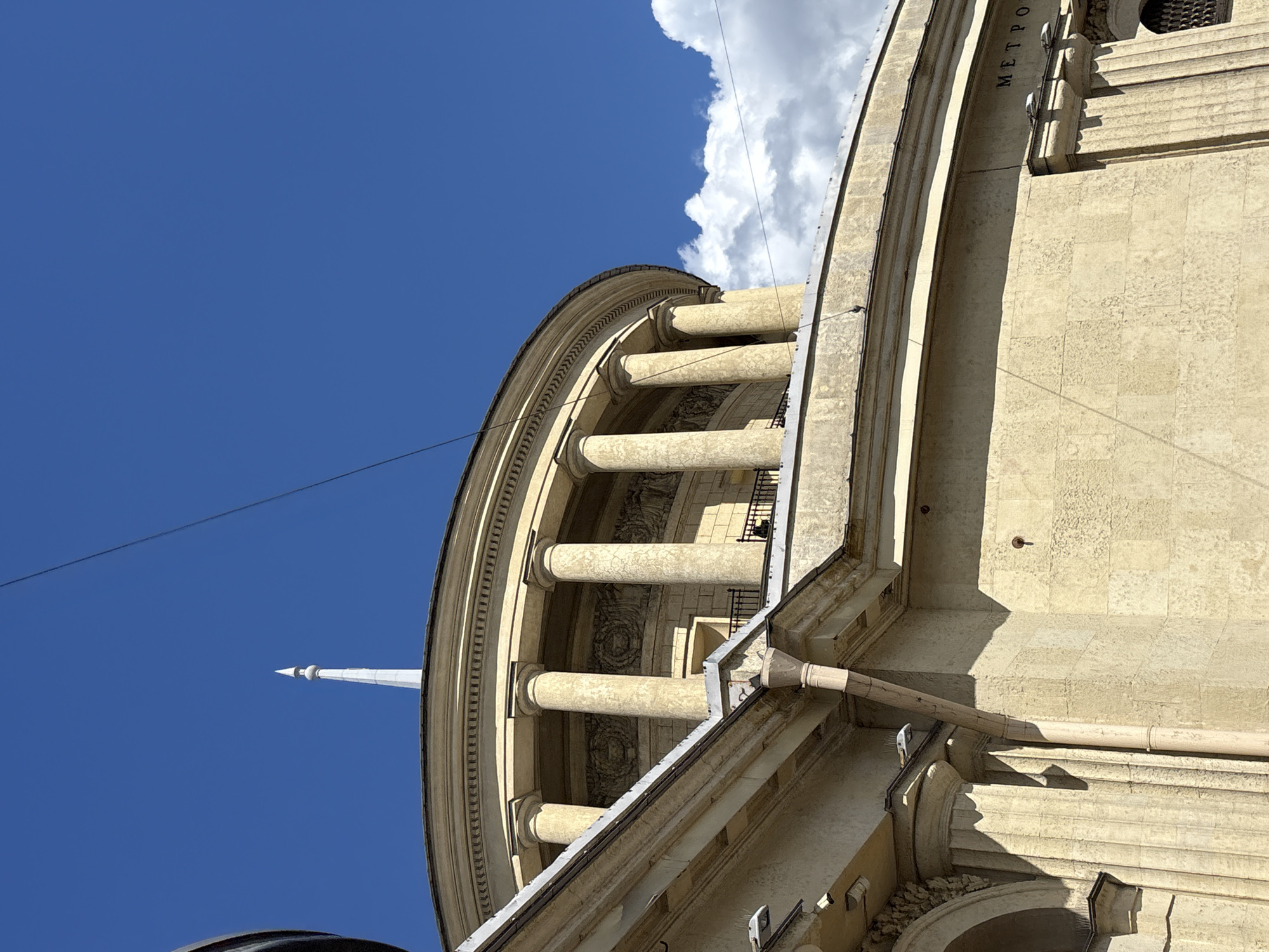 This is a low-angle shot of the Kazan Metro station entrance in Saint Petersburg, Russia, on a sunny day with a bright blue sky and scattered clouds. The main subject is the grand, circular building with classical architecture, featuring a prominent dome topped by a spire. The building is constructed of light-colored stone blocks and is adorned with a colonnade of tall, fluted columns. Decorative friezes are visible between the columns. A rainwater pipe runs down the side of the building, casting a shadow. The word "METPO" (likely the beginning of "METRO") is inscribed in black letters on the building's facade. A thin wire stretches across the sky above the building.