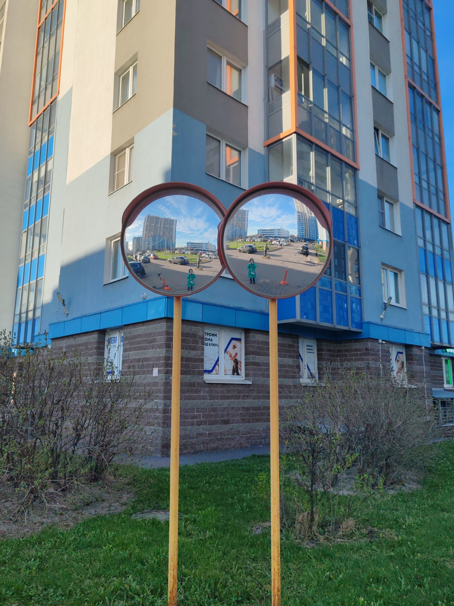 Two convex safety mirrors on yellow poles stand in a grassy area with bushes, reflecting a scene across the street. A person in a green coat stands near a traffic cone in front of a building with blue accents. Cars and a scooter are visible in the reflection. The building in the background has a modern design with a combination of light and dark gray, white, and orange details. The building's lower portion is blue. Posters are affixed to a brick-faced building section in the foreground. The scene is situated in Saint Petersburg, Russia.