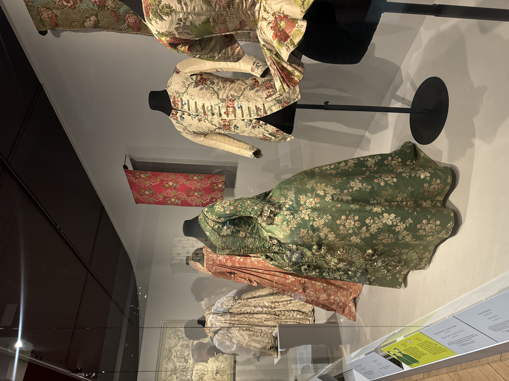 The image depicts a display of historical clothing, likely in a museum setting. The clothing consists of several dresses and a jacket, all with elaborate floral patterns and designs. The clothing is arranged on mannequins, some of which are mounted on stands. The dresses range in color from green to peach to a light, patterned color. A jacket with a lighter background color and detailed floral accents is also showcased. In the background, a patterned textile piece hangs on the wall. To the right side of the image, descriptive cards or plaques can be seen, providing information about the displayed items. The setting appears to be a well-lit exhibit case. The location may be in Paris, France.