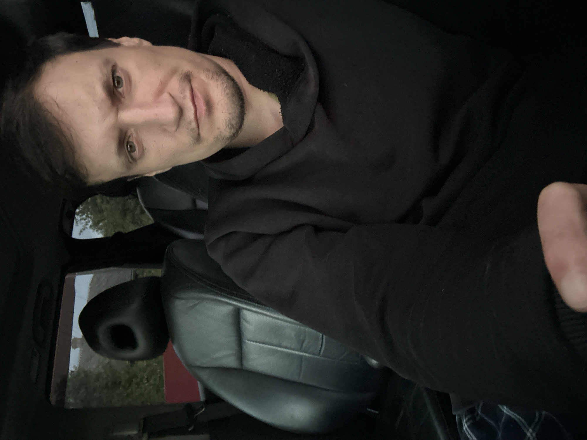 A man, appearing to be in his late 20s or early 30s, is seated in the driver's seat of a car. He is wearing a black hoodie and has short, dark hair. His gaze is directed towards the camera, and he has a faint smile on his face. The interior of the car is visible, including black leather seats and a headrest with a distinctive circular cutout. Outside the car, trees and a red fence can be seen through the windows, suggesting a rural or suburban setting. The lighting indicates it is either early morning or late afternoon. The image was taken in Makiv, Ukraine.