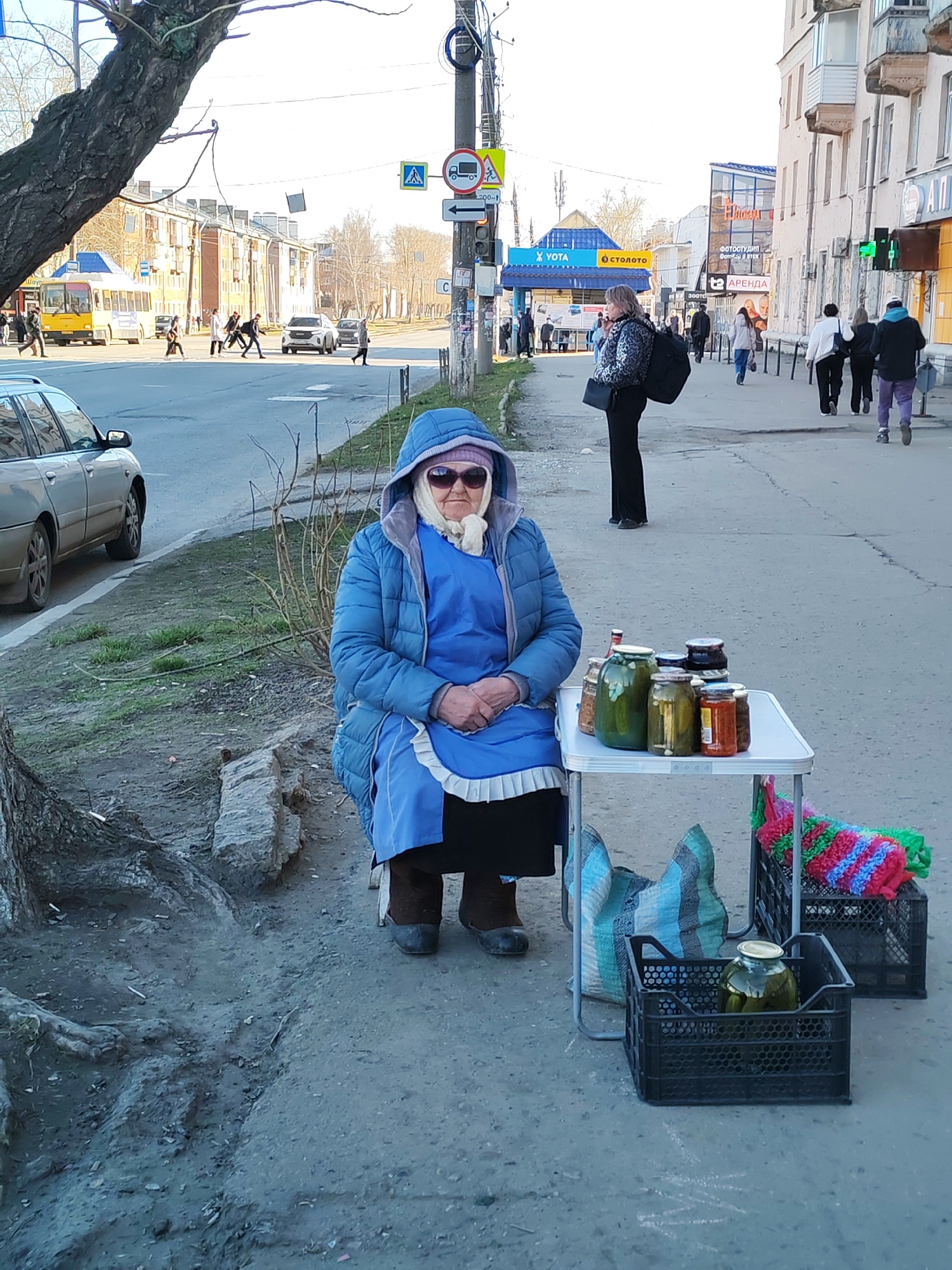 An elderly woman sits on a stool by a dusty city street, engaged in informal vending. She wears a blue puffer jacket with a hood, a blue apron, a white headscarf, and sunglasses. Her hands are clasped in front of her. On a small white folding table beside her, several glass jars of pickled vegetables, including cucumbers, and other preserves are displayed. Below the table, black plastic crates hold additional jars and colorful knitted or crocheted items. A blue and white striped bag rests on the ground near her feet.
To her left, a silver car is parked on the side of the road, and the gnarled roots of a large tree are visible in the immediate foreground. The background shows a bustling city street with pedestrians walking, a yellow bus, and other vehicles. Commercial buildings line the sidewalk on the right. A street light pole with various signs stands in the mid-ground.
Visible text in Cyrillic includes "YOTA" and "СТОЛОТО" on a blue kiosk, and "ФОТОСТУДИЯ" (Photo Studio) and "АРЕНДА" (Rent) on a building. Street signs for a pedestrian crossing, "no trucks," and a letter "C" are also discernible.
The scene takes place on a clear, sunny day in an urban environment, likely in Izhevsk, Russia, as suggested by the location context and Cyrillic script.