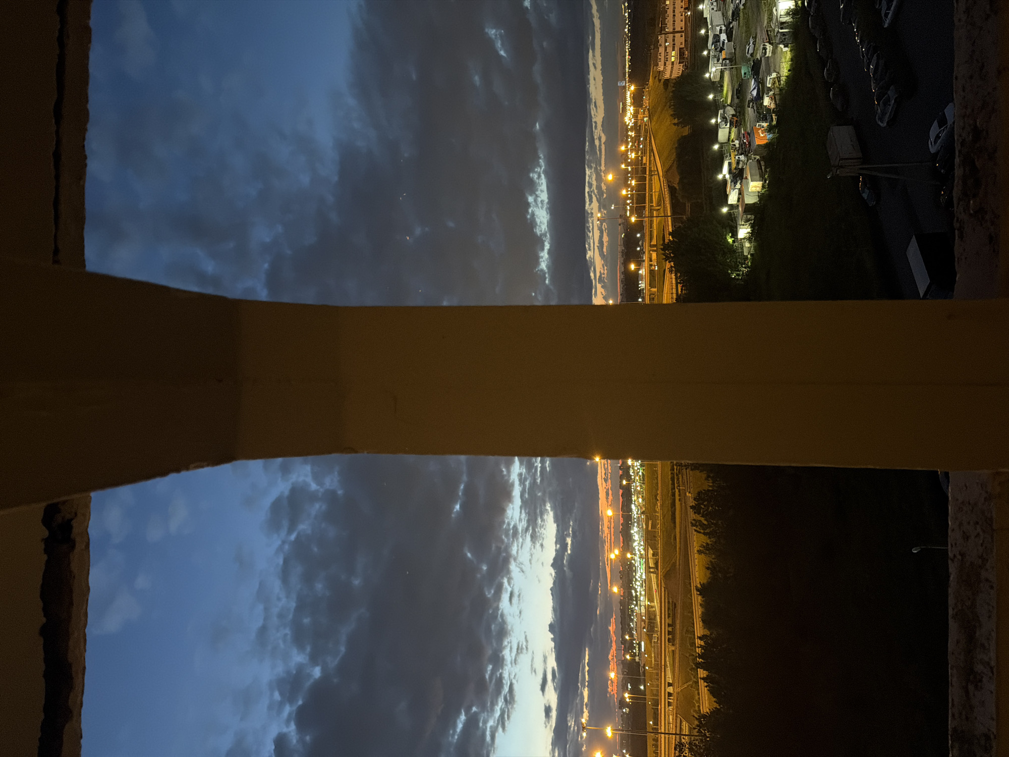 The image shows a view of a city at dusk through a concrete archway. The sky is a deep blue with scattered clouds, tinged with orange and yellow from the setting sun near the horizon. Below the sky, a brightly lit urban landscape is visible, featuring roads, streetlights, and buildings. The foreground is dominated by the dark, textured interior of the concrete structure, with the bottom edge appearing worn and rough. To the left and right of the central archway, glimpses of trees and what appear to be industrial or commercial buildings and a parking lot filled with vehicles can be seen. The overall impression is one of an urban environment transitioning from day to night, viewed from an elevated vantage point.
