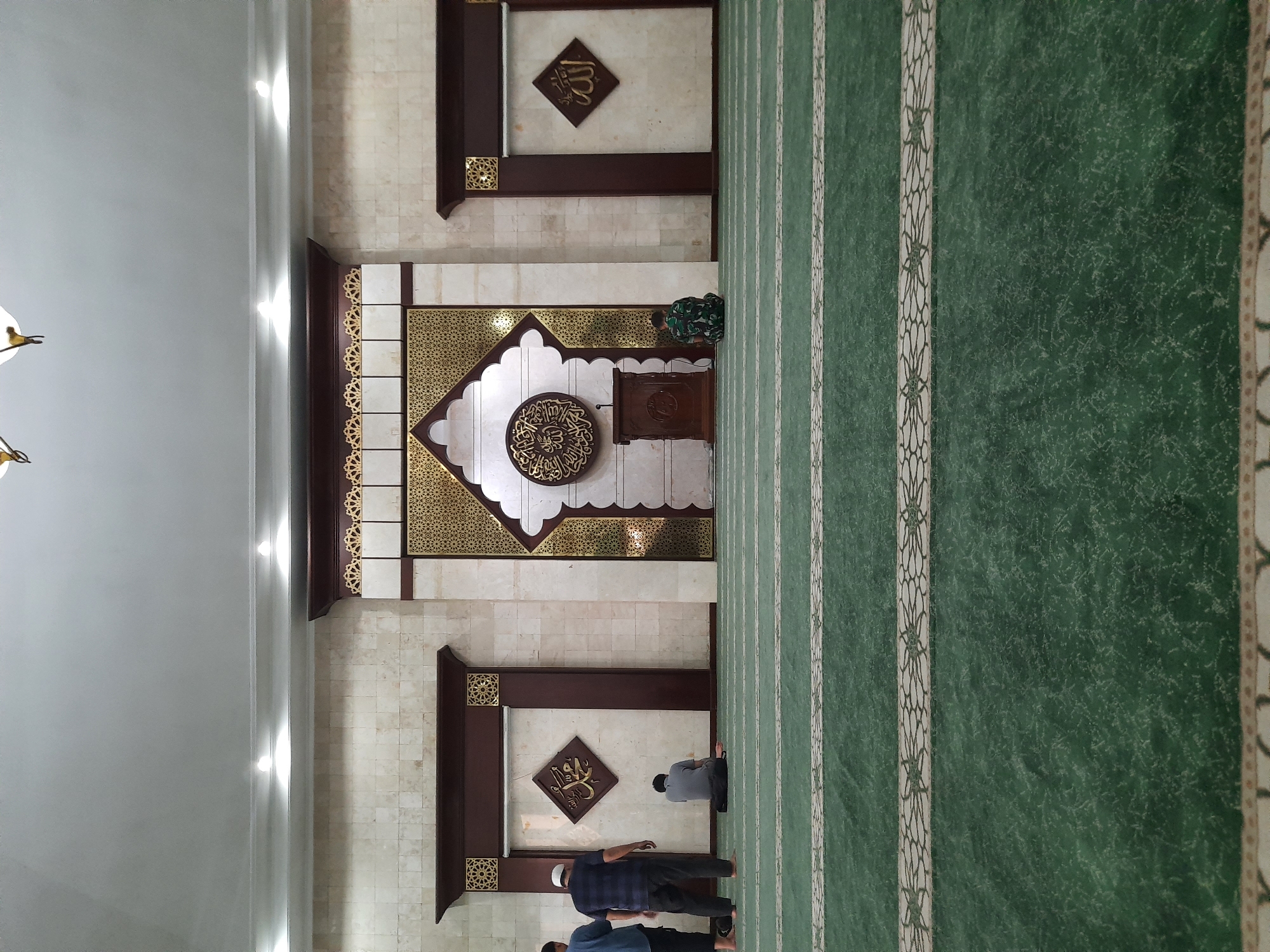 The image depicts the interior of a mosque, likely in Bandung City, Indonesia, characterized by its green carpet with decorative white stripes and a grand, ornate wall. In the foreground, the green carpet covers the floor, featuring two distinct decorative patterns along its width. Three individuals are visible in the mid-ground. On the left, two men stand facing away from the camera, with one wearing a blue shirt and the other a dark blue and white striped shirt, and a white cap. To the right of the standing men, a person in a gray shirt is seated on the carpet, facing the ornate wall. Further to the right, another person, possibly a soldier in camouflage uniform, is also seated on the carpet, facing the same wall.
The focal point of the background is an elaborate wall decoration. At its center, an arched structure is adorned with intricate gold geometric patterns. Within this arch, a circular Islamic calligraphy adorns the wall. To the left and right of this central feature, two framed rhombus-shaped decorative elements are mounted on the tiled walls. The left frame contains gold Arabic calligraphy, and the right frame similarly contains gold Arabic calligraphy. A wooden podium stands in front of the central calligraphy. The walls are tiled with a light-colored stone pattern. The ceiling is white, with recessed lighting fixtures illuminating the space. The overall environment suggests a place of worship, with a sense of order and reverence.