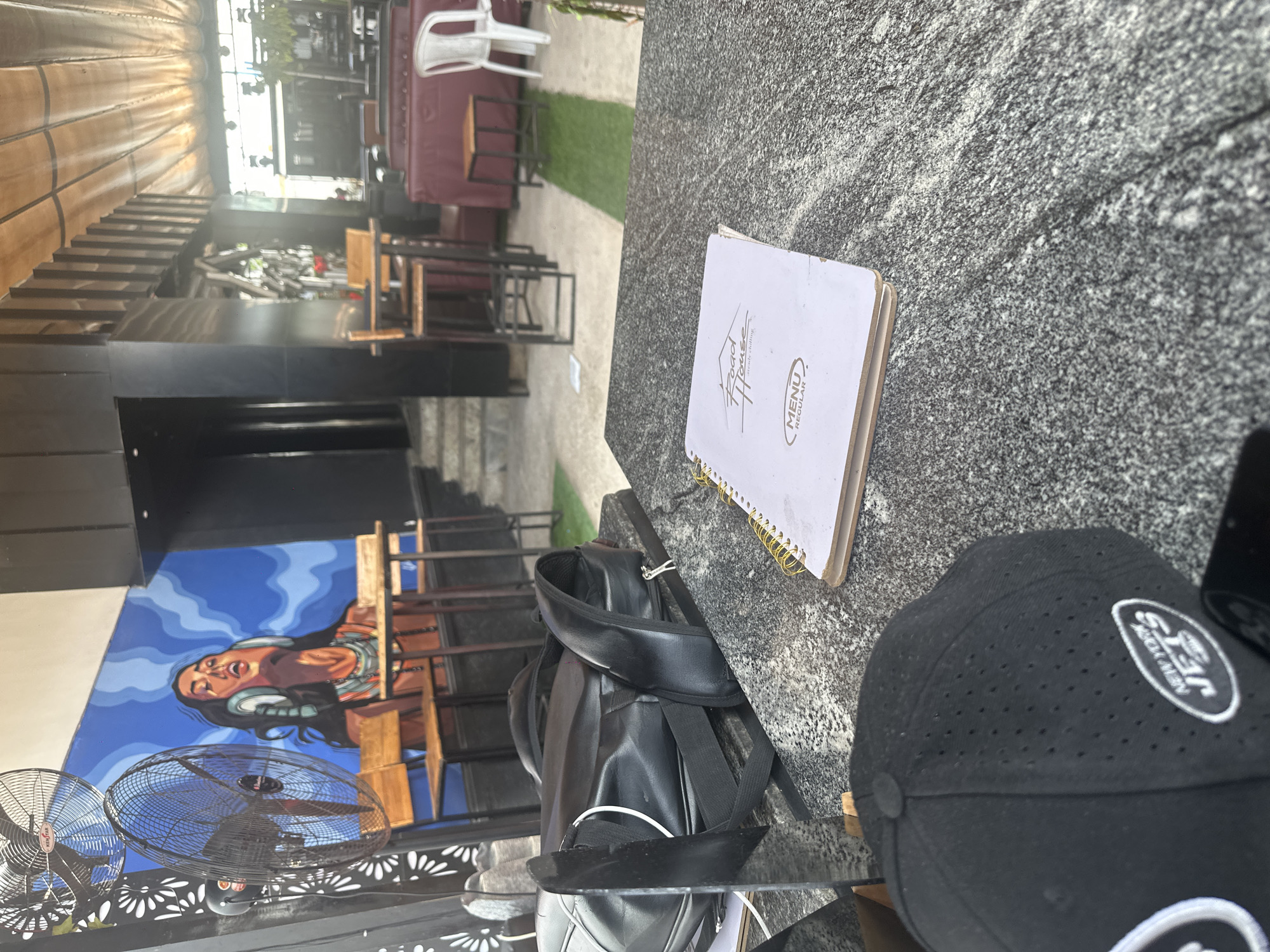 The image captures an outdoor seating area of a establishment, likely a restaurant or bar, in Howayi, Nigeria. The foreground is dominated by a speckled granite table, upon which rests a black baseball cap with "NEW YORK JETS" embroidered on it, and a white menu folder with the text "Road House" and "MENU REGULAR" visible. To the left, a black leather backpack with two straps is placed on the table. The background reveals a lively ambiance with wooden chairs and tables set against a vibrant blue mural of a woman wearing headphones. Two large industrial fans are positioned to the left of the mural. Further back, the establishment features a covered outdoor seating area with maroon booth seating and white chairs. A person is partially visible sitting on a maroon couch in the background. The overall scene suggests a casual, social gathering place with a blend of modern and artistic decor.