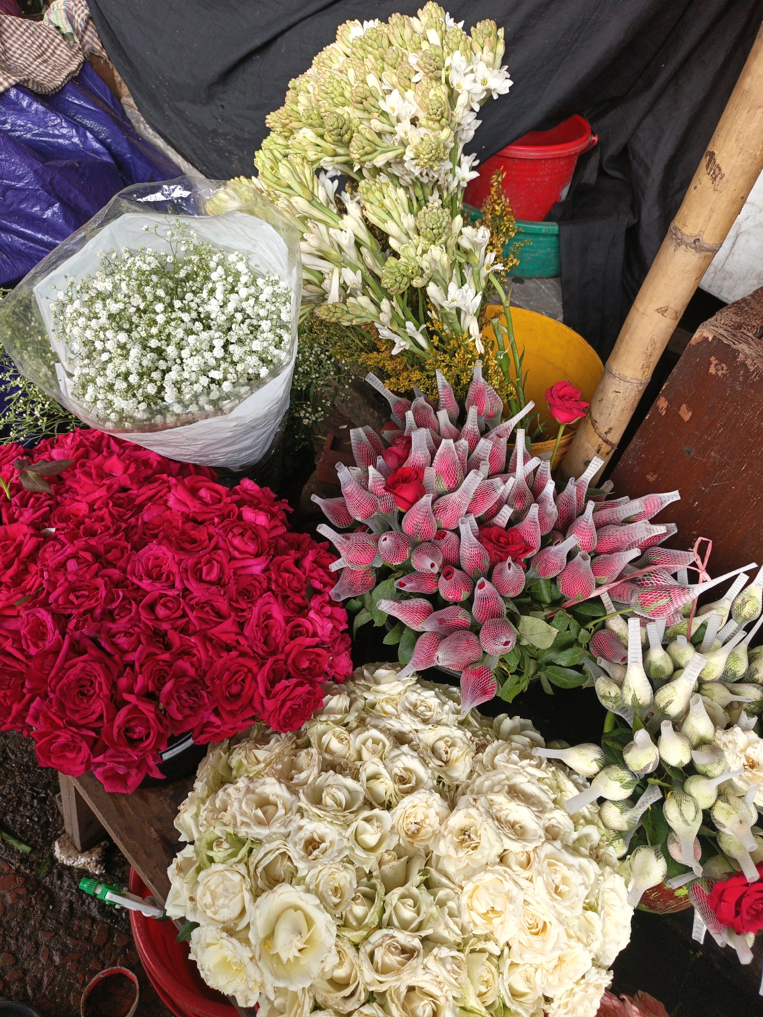 The image shows an assortment of flowers for sale, likely at a market or florist in an unknown city. The flowers include red roses, white roses, white baby's breath, and tuberose. Some of the roses are individually wrapped in netting. A bamboo pole and some containers are also visible in the background.