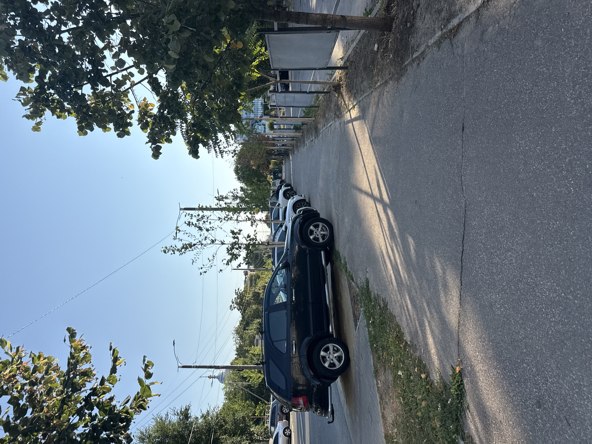 The image depicts a street scene in Chișinău, Moldova. A dark-colored SUV is parked on the right side of a paved road, adjacent to a sidewalk lined with trees. Several other vehicles are parked in the background. The sun is shining, casting shadows on the pavement. Trees with green foliage are present on both sides of the street, with a few buildings visible in the distance. The sky is clear and blue.