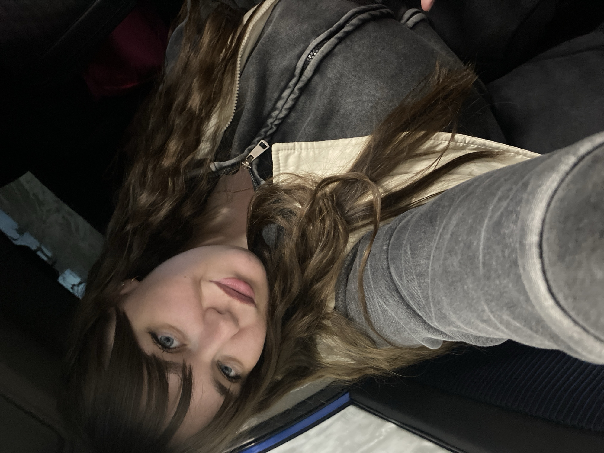 A woman with long, wavy brown hair and blue eyes is facing the camera. She is wearing a grey, hooded jacket with a white collar. The setting appears to be inside a vehicle. The background is dark, with some indistinct shapes. The image is rotated. There is no information to suggest a location in the city of Votkinsk, Russia.