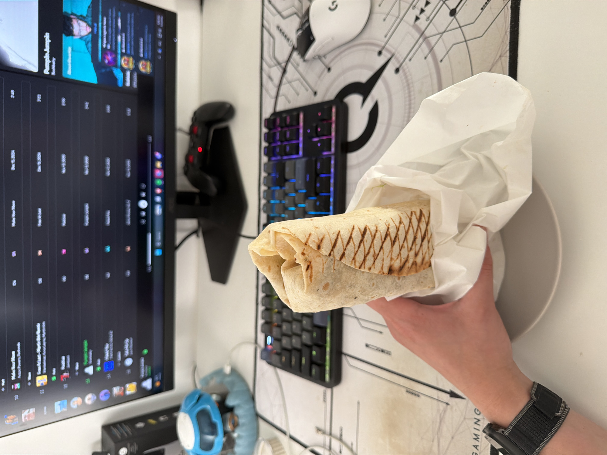 A person's hand holds a wrapped shawarma in front of a computer setup. The computer screen displays a playlist, and a gaming mouse and keyboard are visible. The background includes a gaming mousepad with a logo. The environment appears to be indoors, likely a home or office setting. There's no information that indicate location in Belgrade, Serbia.