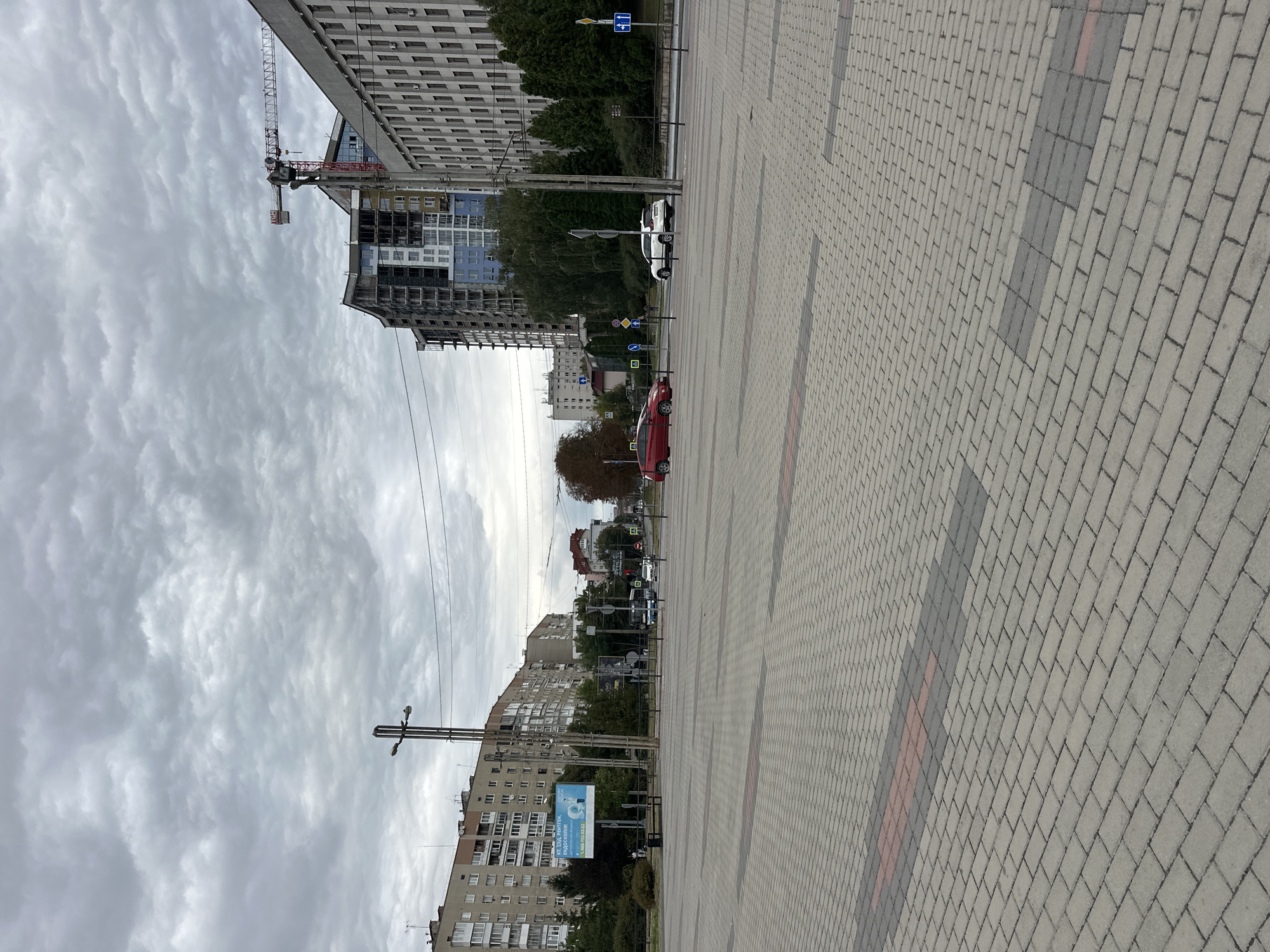 The image depicts an overcast day in Ternopil, Ukraine, showcasing a wide, paved pedestrian area in the foreground with a decorative pattern of grey and red rectangular pavers. Beyond the paved area, a street runs horizontally, with traffic visible. On the left side of the street, there are several multi-story residential buildings and a billboard advertising medical services. A street lamp stands prominently in the mid-ground. Across the street, on the right, a modern building under construction with a crane is visible, adjacent to a larger, older, multi-story administrative-style building. Green trees line the street between the buildings. The sky is filled with dense, grey clouds.