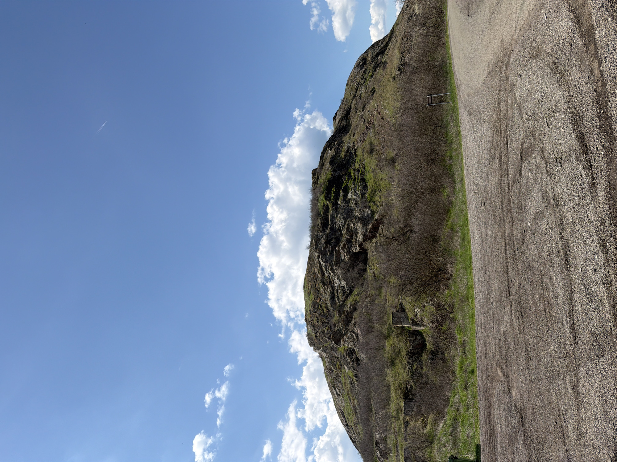 The image shows a rocky cliff face next to a gravel road under a blue sky with scattered clouds. A faint contrail stretches across the upper left portion of the sky. Green vegetation clings to the cliff and grows along the roadside. A short metal railing stands near the base of the cliff. The location is near Rusokastro, Bulgaria.