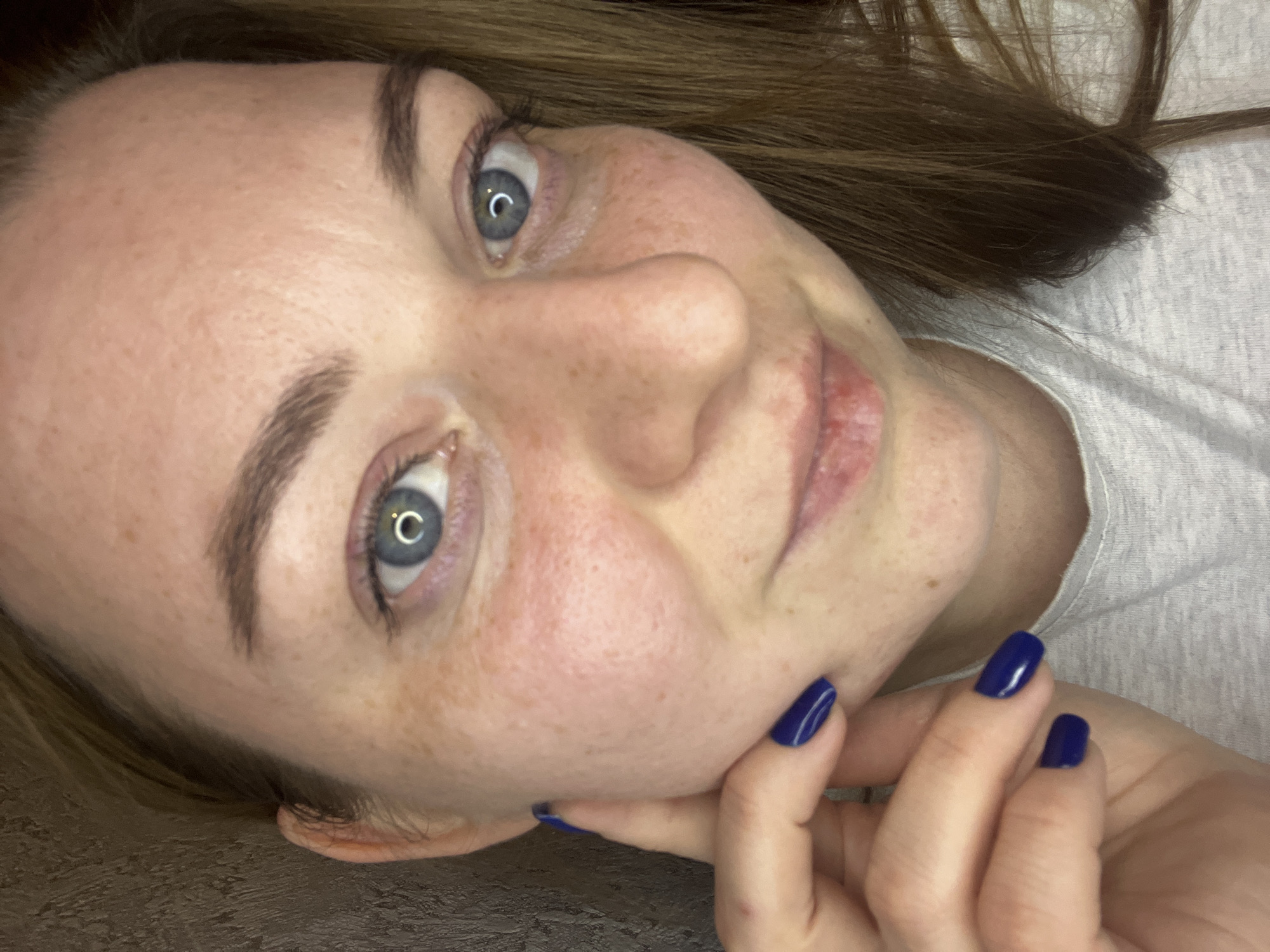 A fair-skinned woman with light brown hair is positioned close to the camera. Her face is tilted slightly. She has blue eyes, freckles, and well-defined eyebrows. Her left hand is near her face. Her fingernails are painted a dark blue color. She appears to be wearing a light-colored t-shirt. The background is blurry and does not depict Раздолье, Russia.