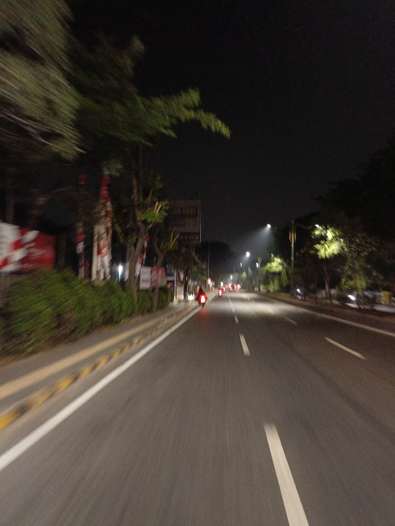This is a night-time, first-person perspective shot of a street in Wangunharja, Indonesia. The camera is in motion, likely on a motorcycle, indicated by the motion blur on the left side of the image showing trees and foliage, as well as red and white banners. The road ahead is paved and has white lane markings. Streetlights illuminate the scene, with some trees visible on the right side of the road. Several motorcycles with red taillights are seen in the distance, suggesting traffic. The overall impression is of a busy street at night.